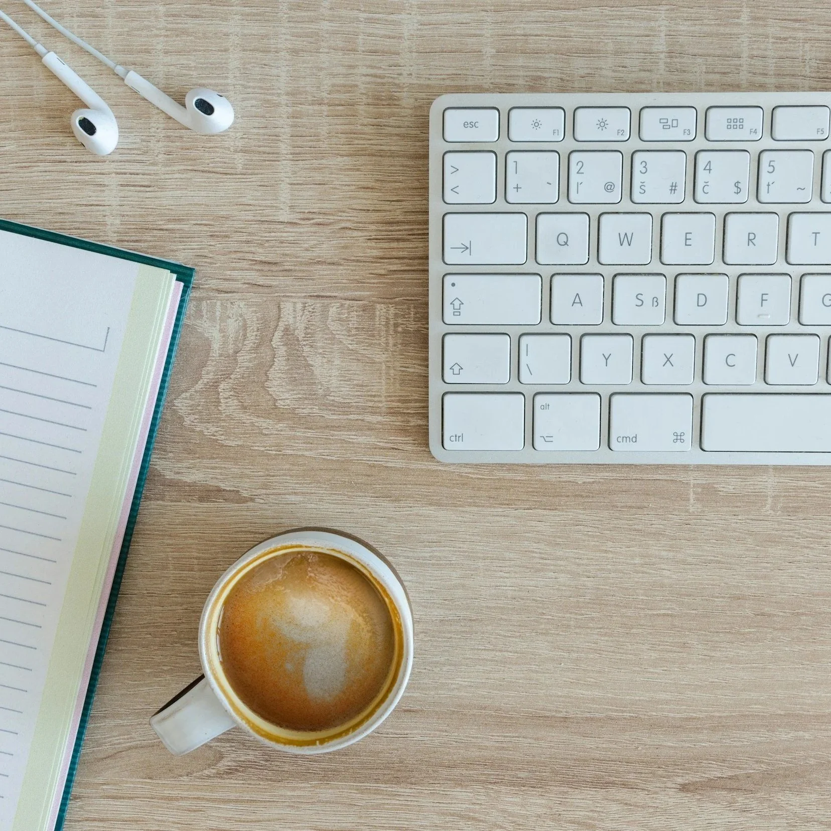 A top-down view of a workspace with a coffee mug, a white keyboard, a notebook, and a pair of earphones on a wooden surface. Representing organized workspace for improved flow.