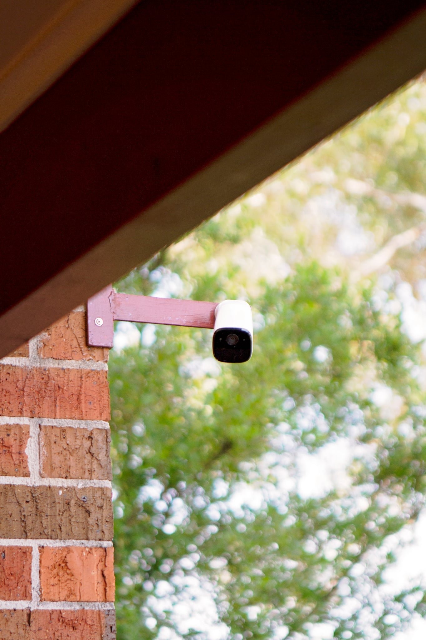 A security camera mounted under a brick archway outside a building, with trees and sky in the background.