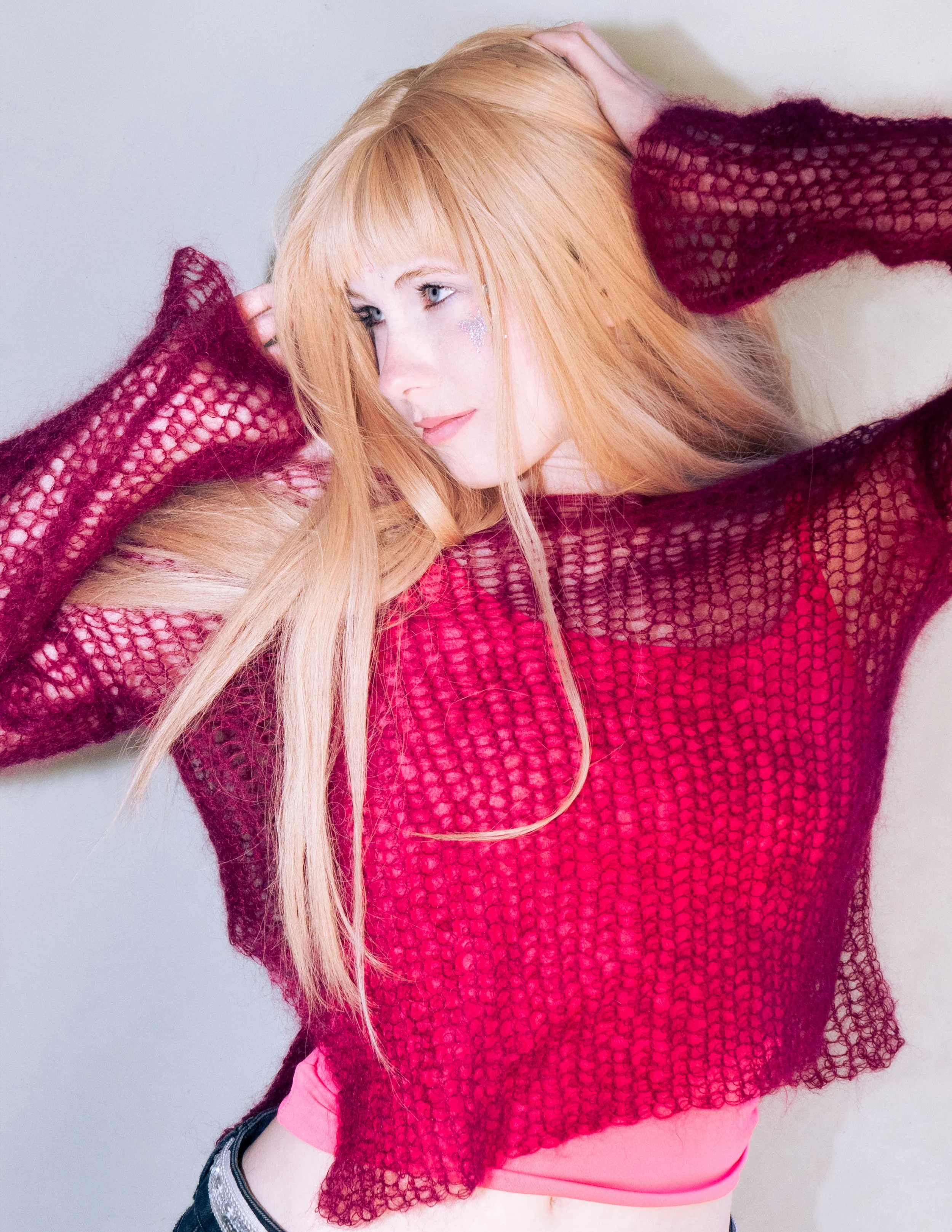 A young woman with long, blonde hair and fair skin, wearing a red mesh top and pink undergarments, posing with her hands on her head against a plain, light background.