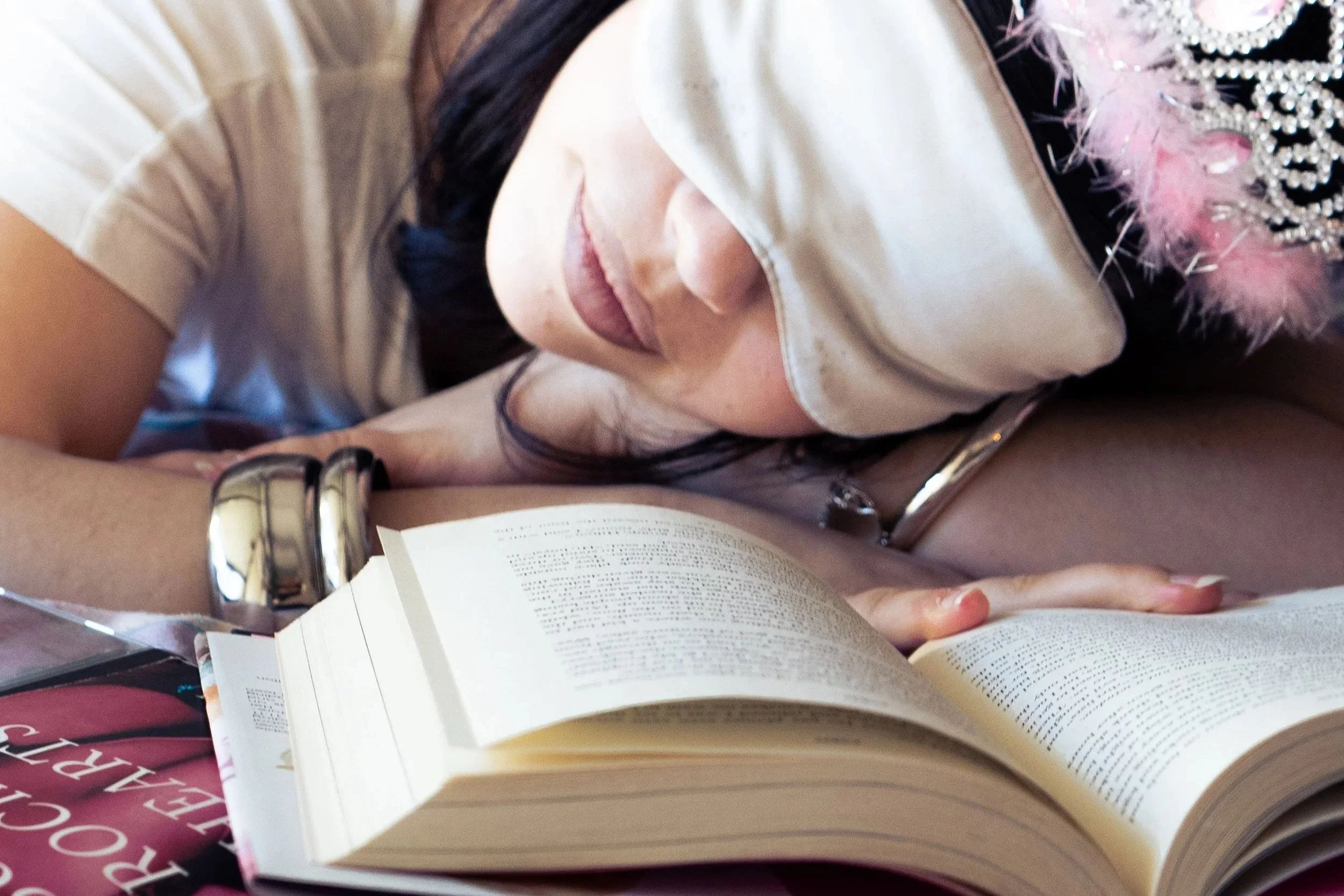 A person reading an open book with visible text, wearing a silver watch and a bracelet. The person is also wearing a white top, and the background contains a pink and purple fuzzy item decorated with pearls and rhinestones.