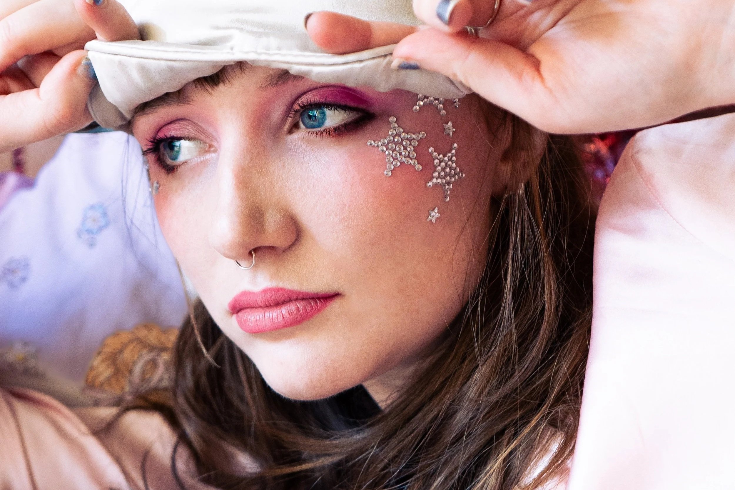 A young woman with bright blue eyes and long brown hair, putting on a white shirt, with star-shaped rhinestone face gems on her cheek and pink eyeshadow.