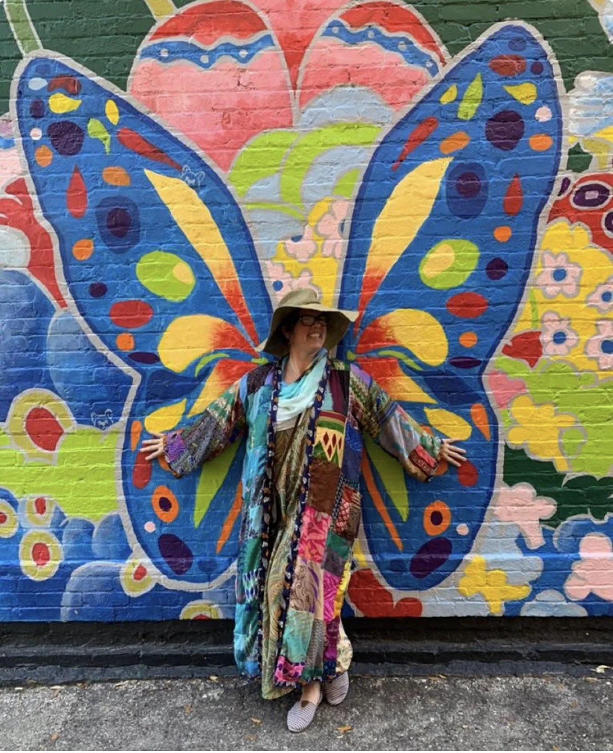 A person smiling and posing with arms outstretched in front of a colorful butterfly mural painted on a brick wall, wearing a wide-brimmed hat, glasses, and a patchwork coat with various patterns and colors.