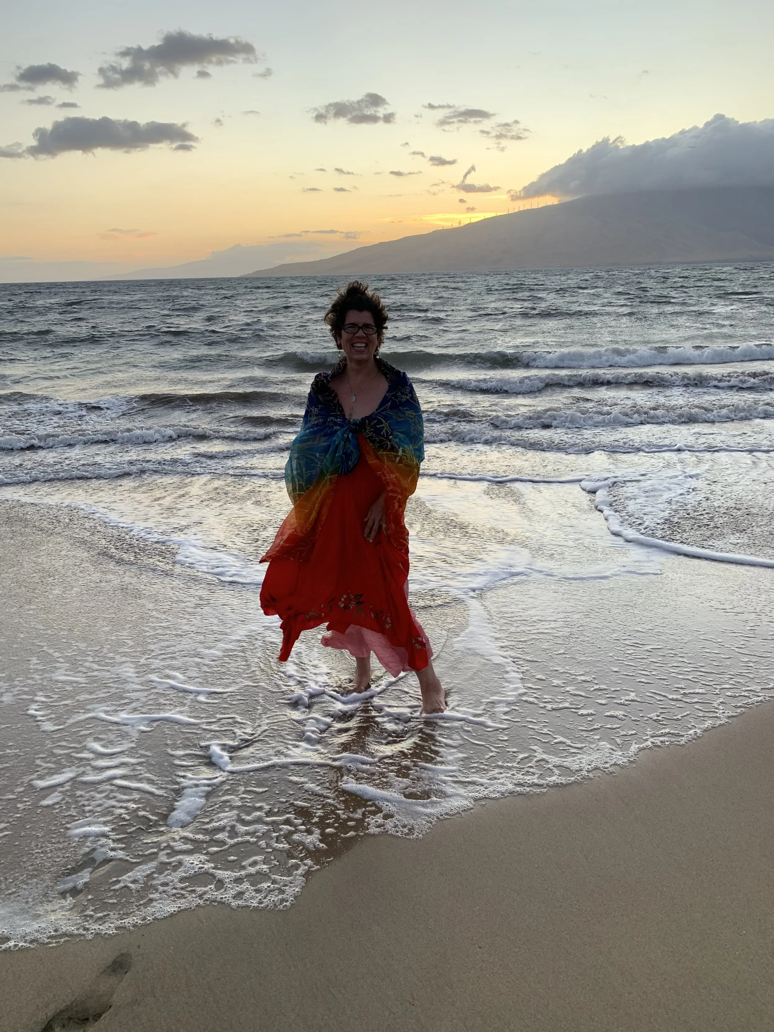 A woman wearing glasses, smiling, and wrapped in a colorful shawl, standing at the edge of the ocean with small waves and a mountain in the background during sunset.