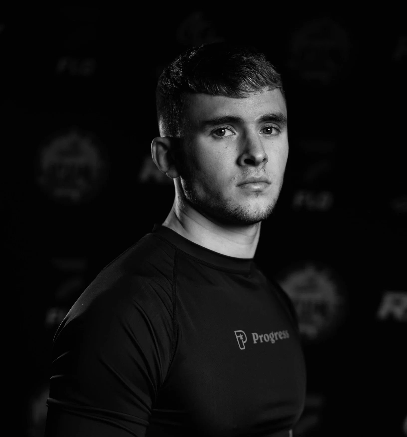 Black and white portrait of a young man with short hair wearing a sports shirt with the logo 'Progress' on it, looking directly at the camera.