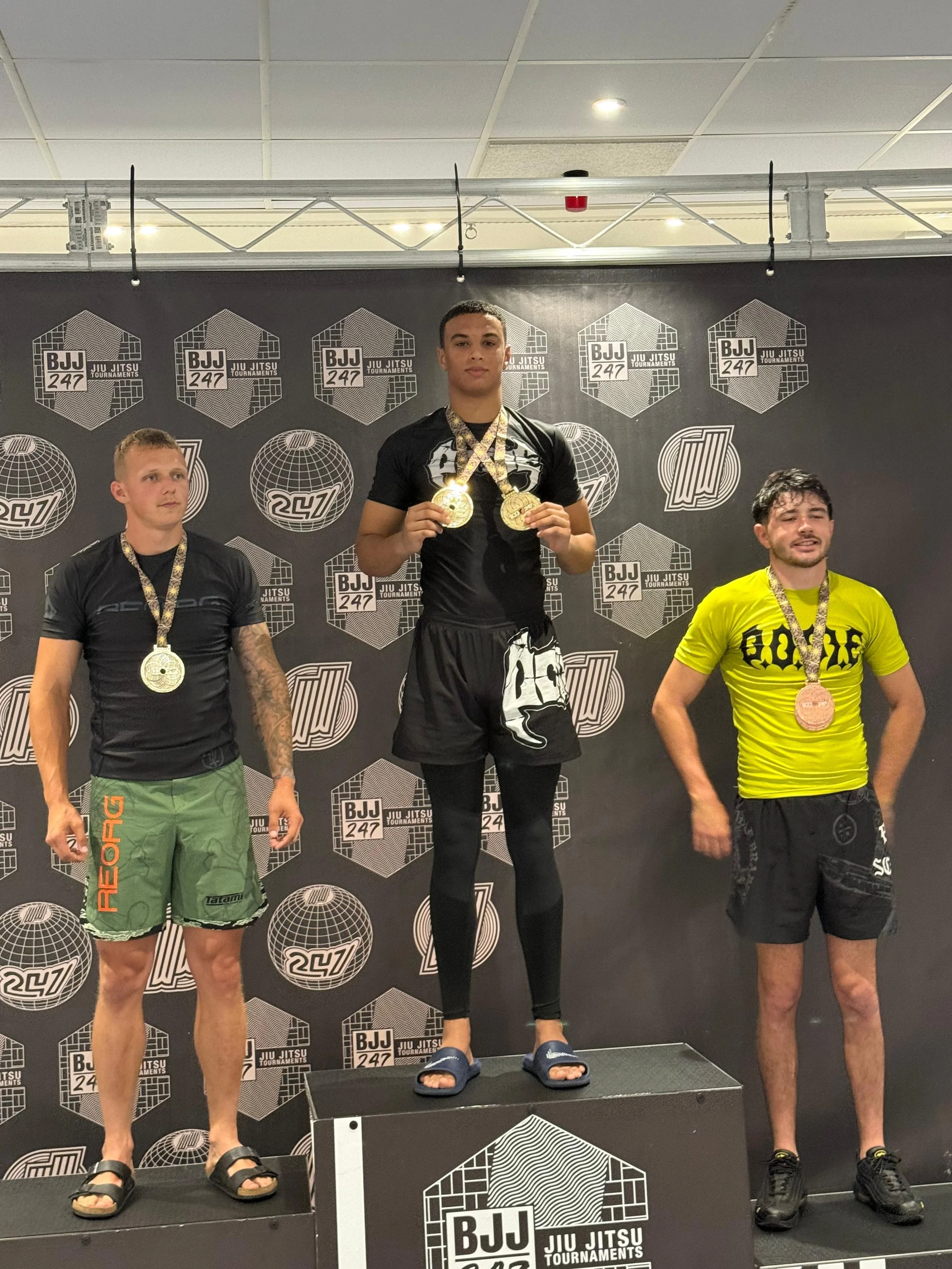 Three male athletes standing on a winners' podium, each with medals around their necks, in front of a backdrop with logos from the BJJ 247 Jiu Jitsu tournaments, celebrating their victory.