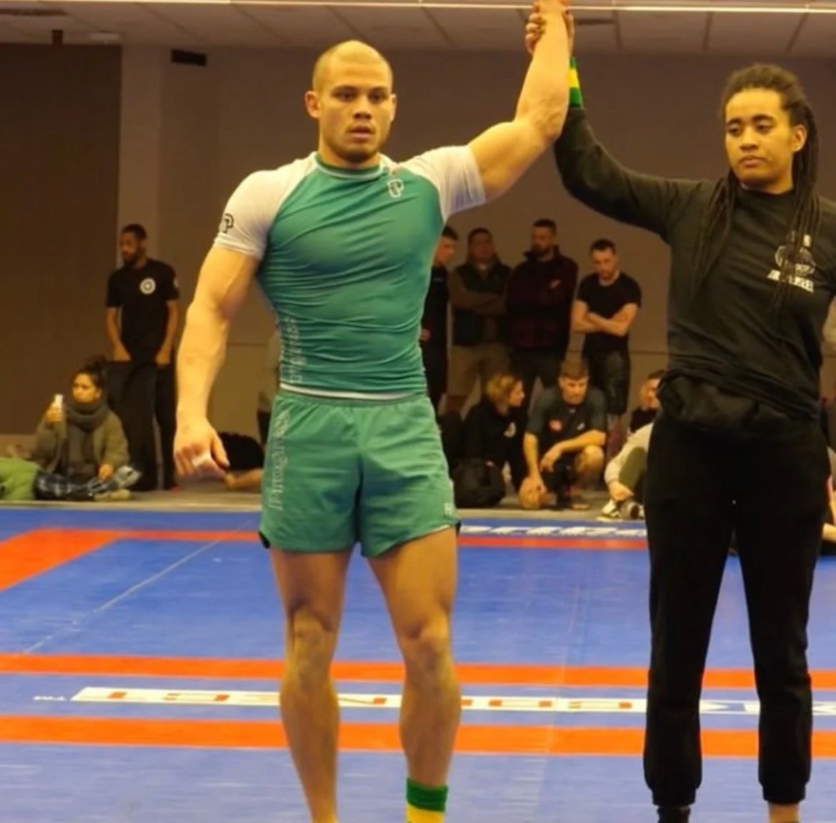 A male athlete in green sportswear raising his arm, with a referee in black holding his wrist, inside a gymnasium with spectators in the background.