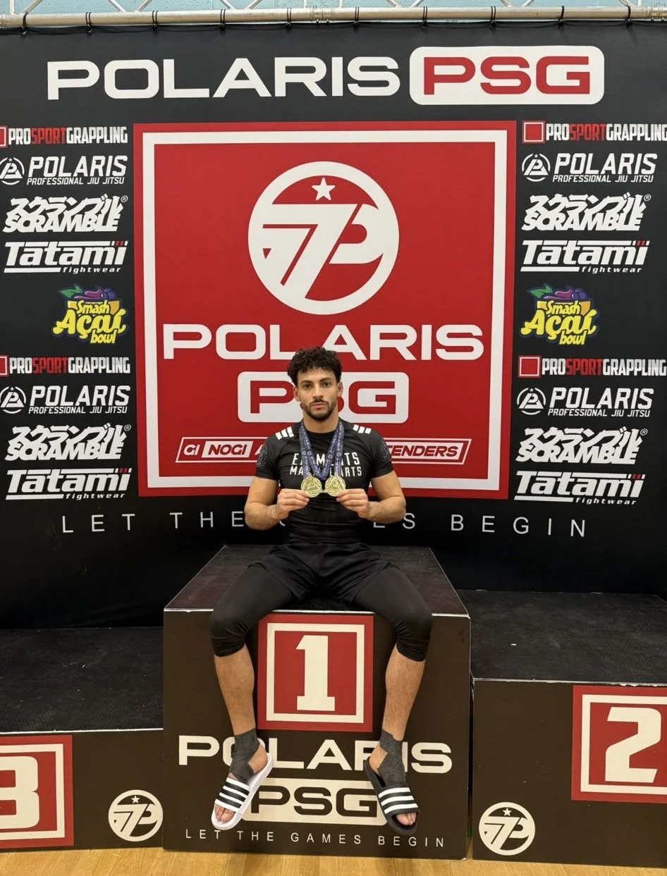 A young man sits on a winners' podium, holding gold medals. He wears a black sports shirt and shorts, and black slide sandals with white stripes. Behind him is a red and white banner displaying the logos of Polaris PSG and other sponsors, with the slogan 'Let the Games Begin'.
