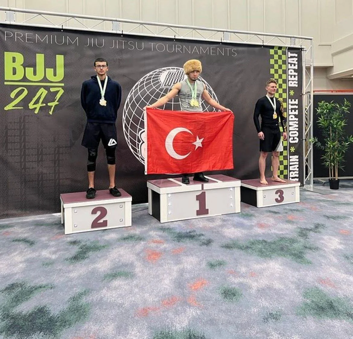 Three male competitors on a winners' podium at a BJJ tournament, with the first-place winner holding a Turkish flag and wearing a fur hat, medals around their neck, and standing in front of a banner that reads 'PREMIUM JIU JITSU TOURNAMENTS'.