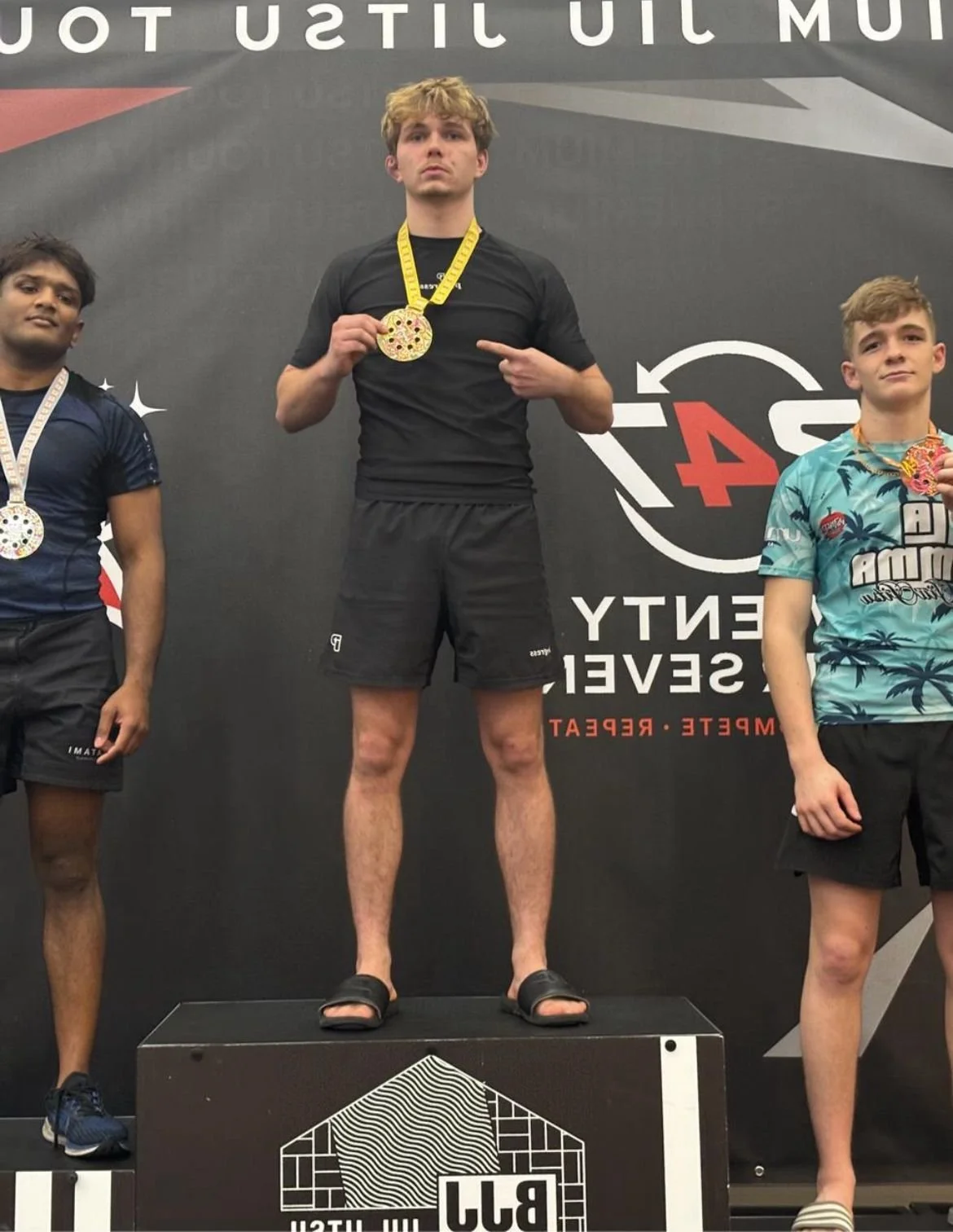 Three young male athletes standing on a winners' podium, each wearing medals around their necks, at a sports event with a black backdrop featuring event branding.