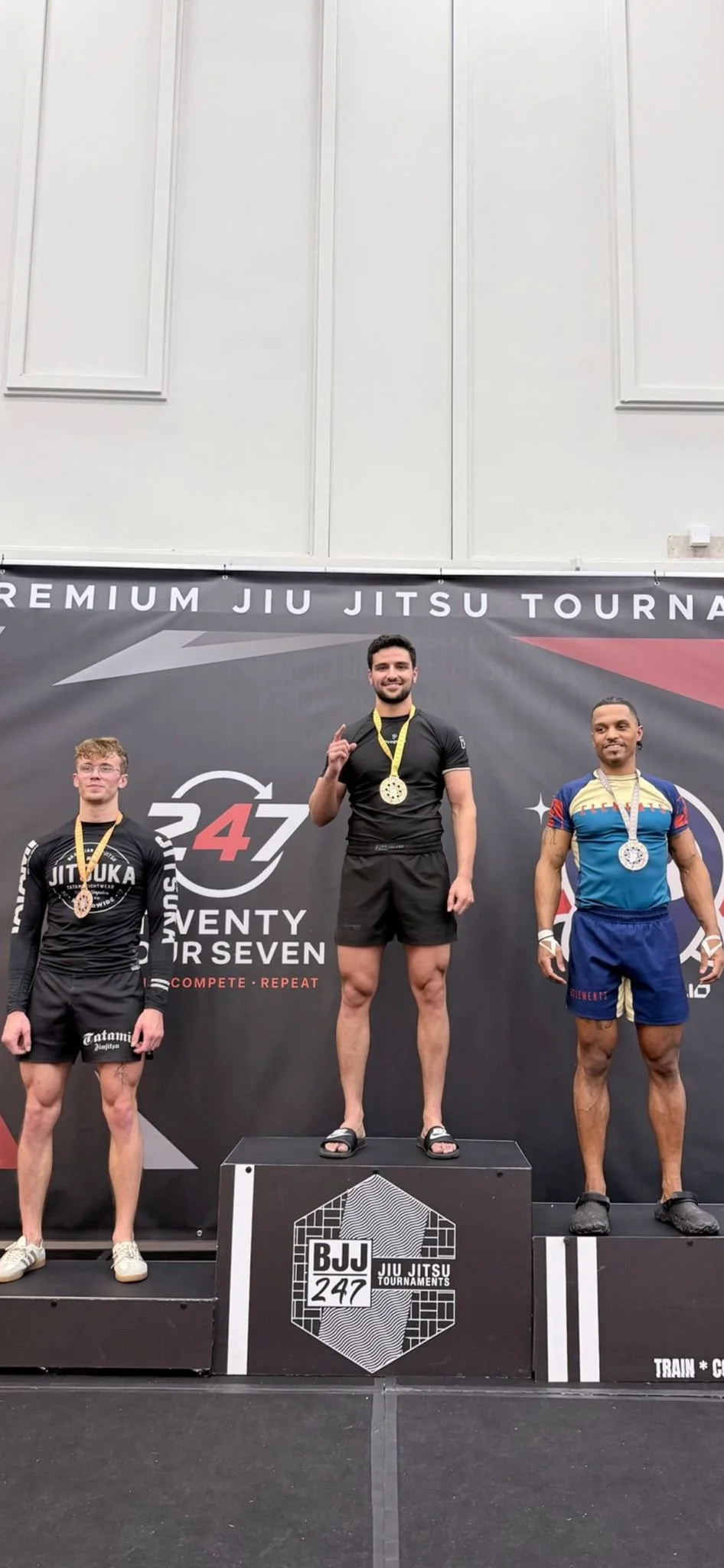 Three male athletes standing on a winners' podium at a Jiu Jitsu tournament, with medals around their necks. The central athlete is on the top step, smiling and making a hand gesture. The background features banners with event branding.
