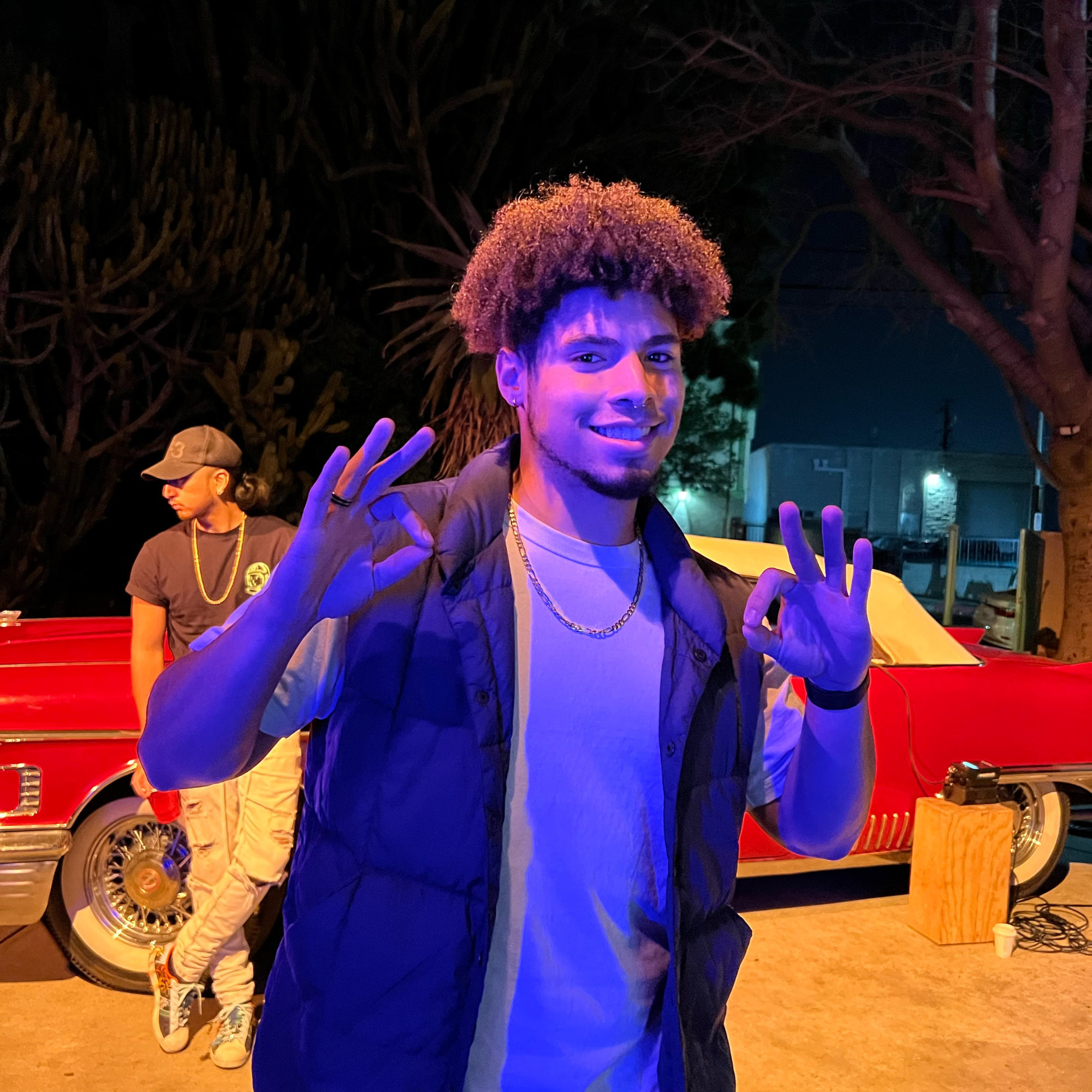 A young man with curly hair smiling and making an OK hand gesture with both hands at night, with a vintage red car and a woman in the background.