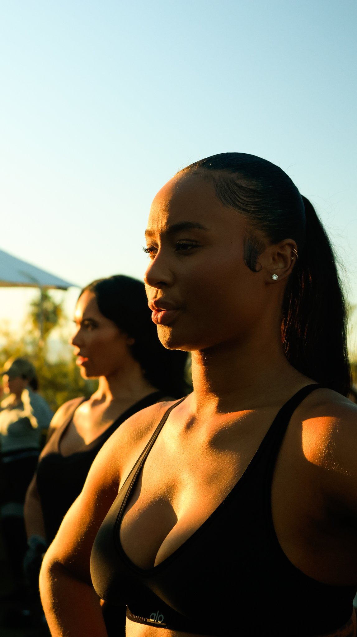 Two women wearing black tops, standing outdoors during sunset, with a blurred background of trees and sky.