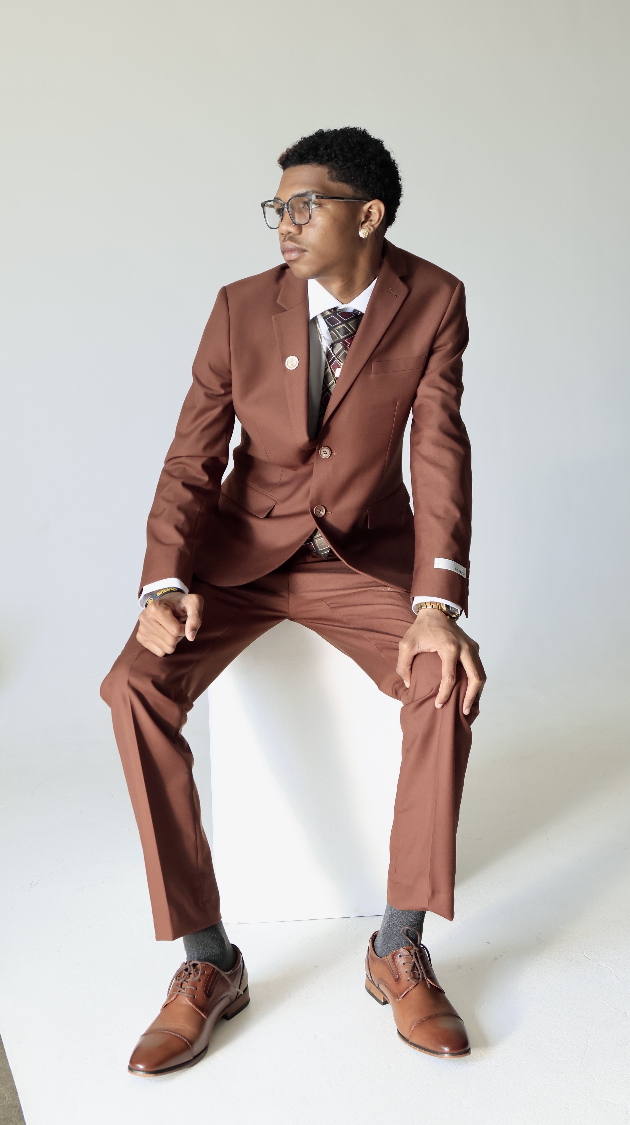 A man in a brown suit, white shirt, patterned tie, gray socks, and brown dress shoes, posing with a serious expression against a plain background.