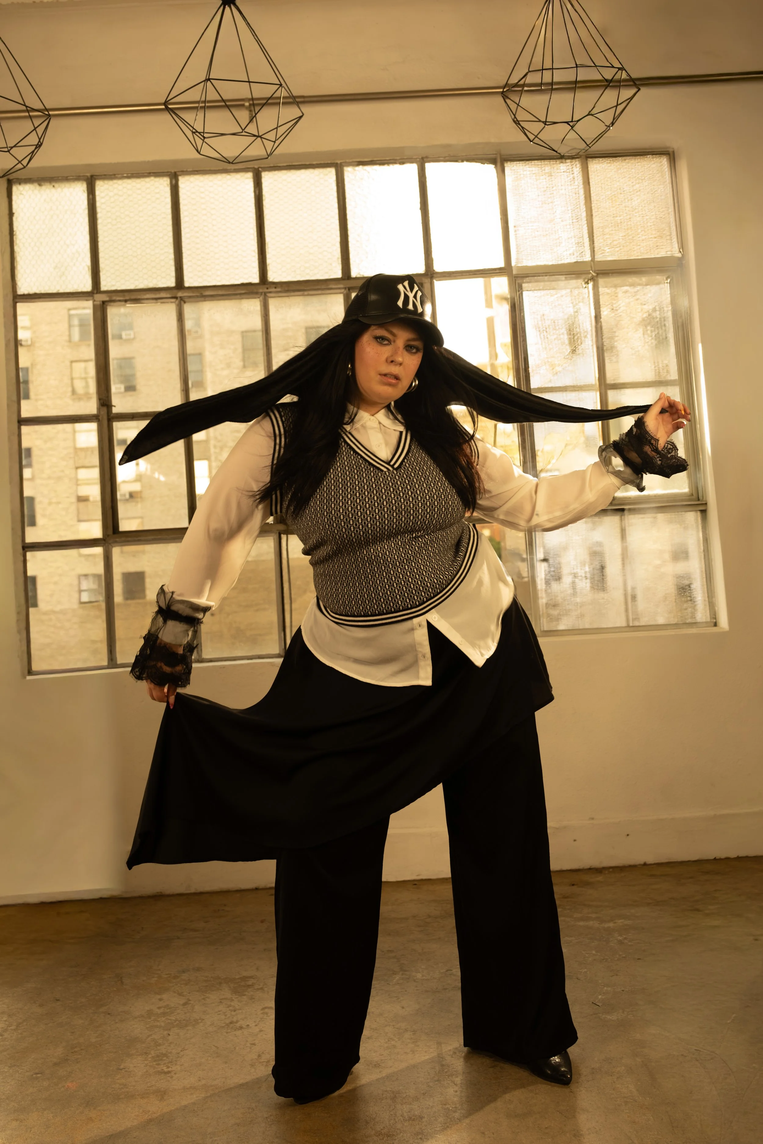 A woman in streetwear posing in an industrial-style room with large windows and exposed pipes, wearing a New York Yankees cap, a patterned vest, a white blouse, black wide-leg pants, and lace cuffs.