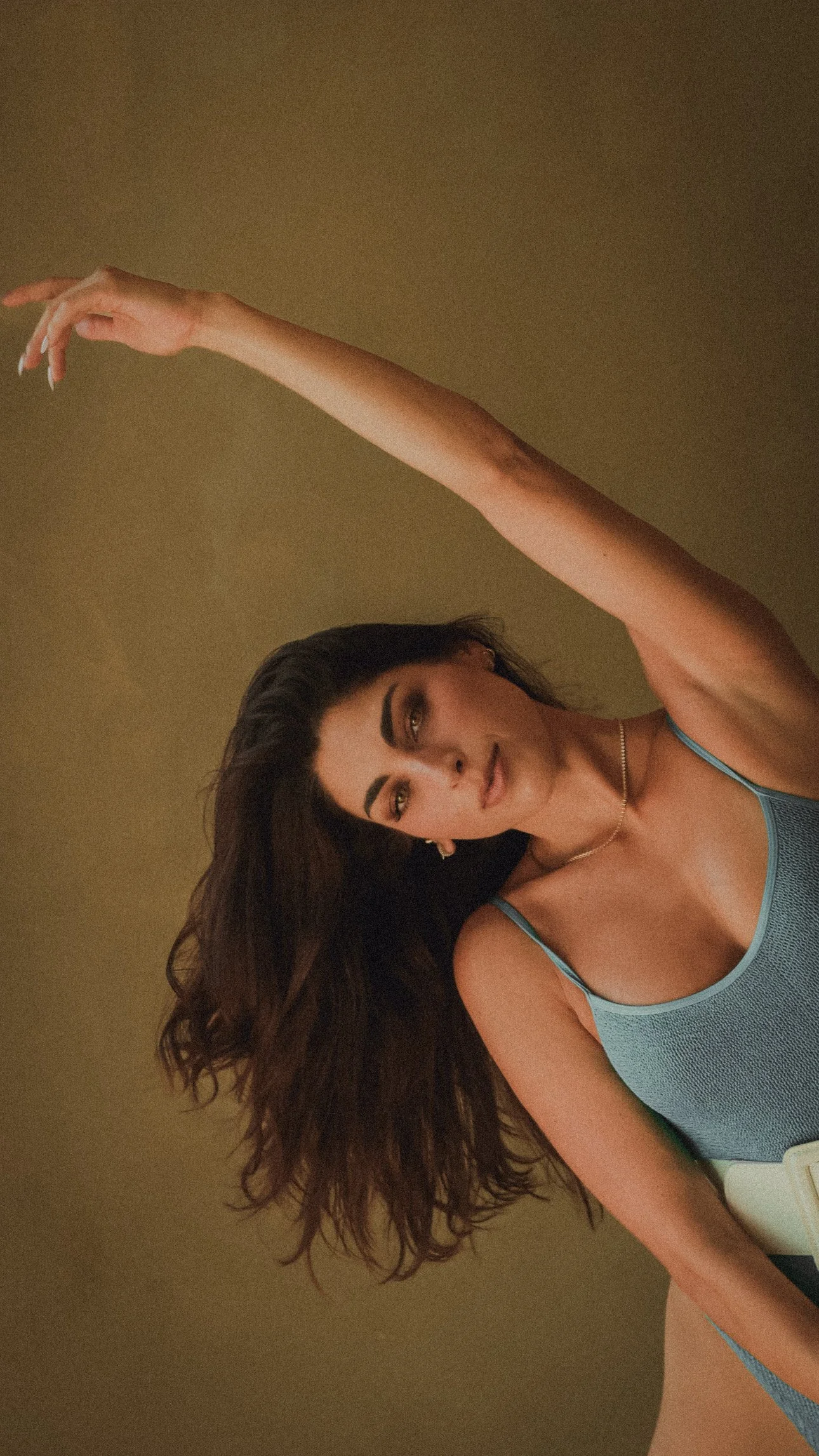 A woman with dark hair and makeup, wearing a blue tank top and jewelry, posing with her arm extended above her head against a plain background.