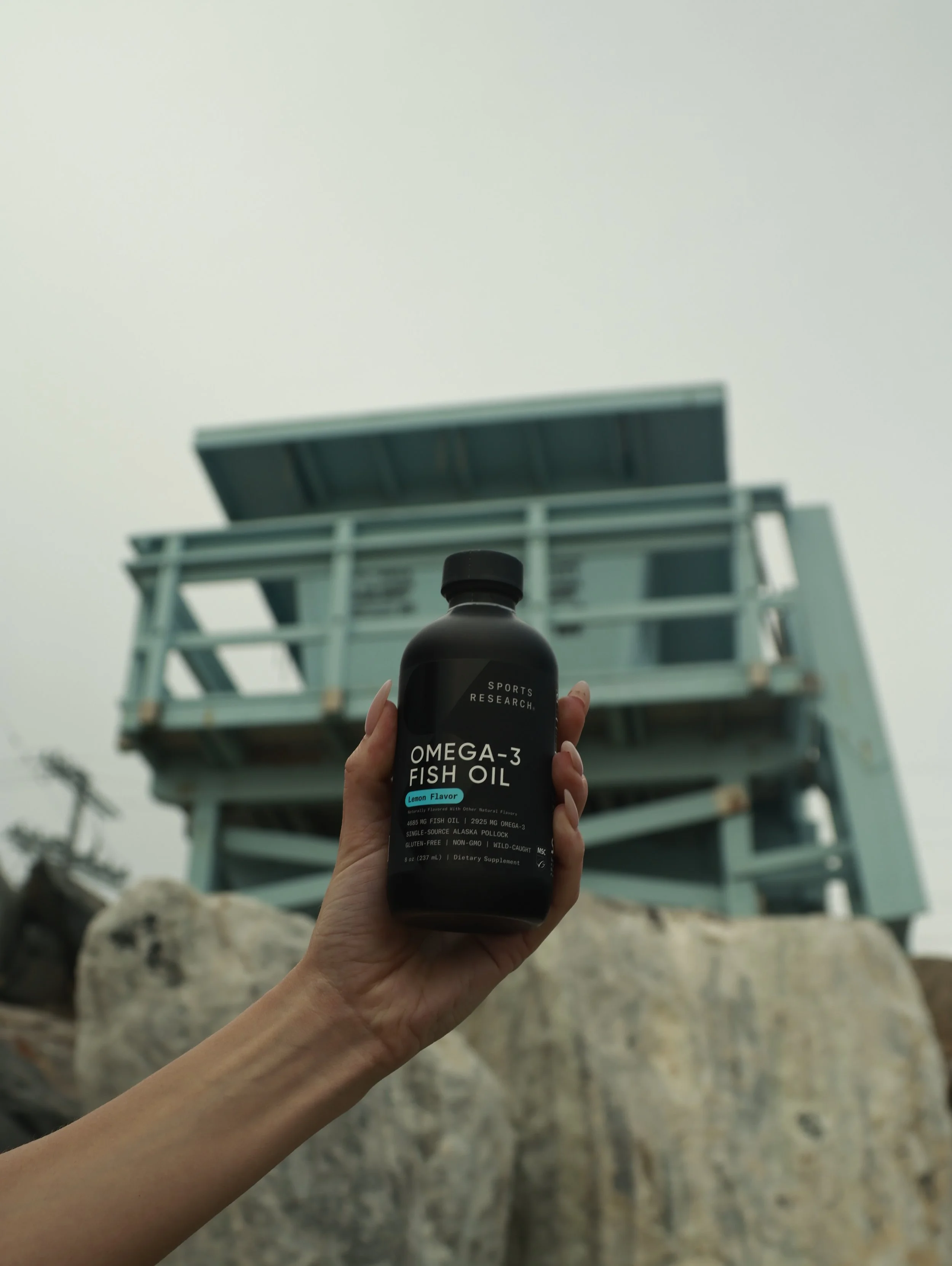 A hand holding a bottle of Omega-3 Fish Oil supplement in front of a lifeguard tower on a beach.