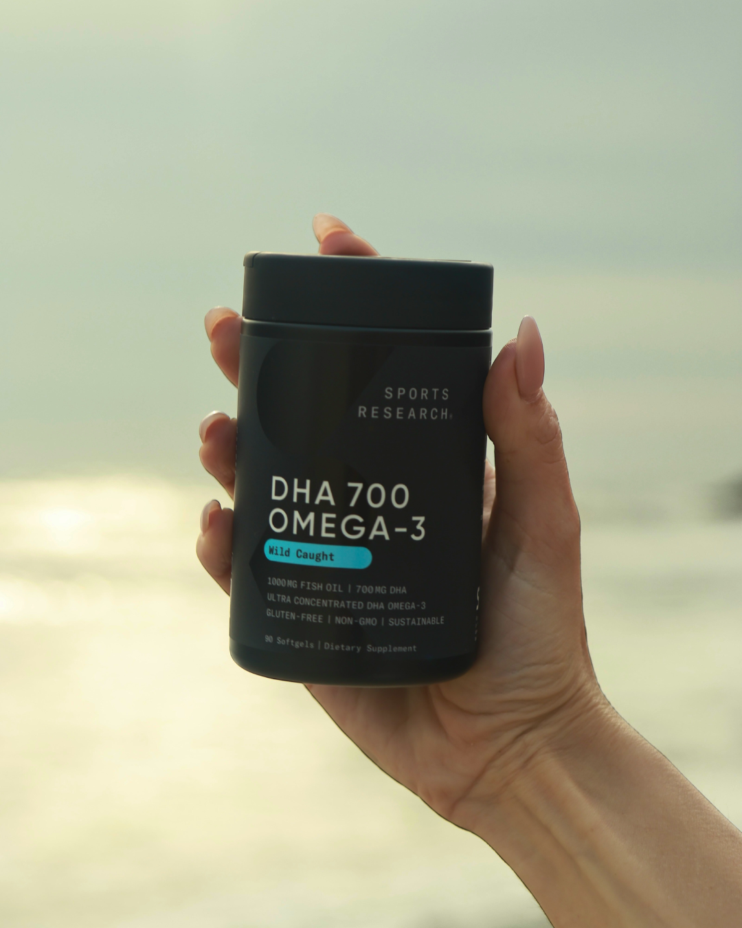 A hand holding a black supplement container labeled DHA 700 Omega-3 with a background of a body of water and sky.