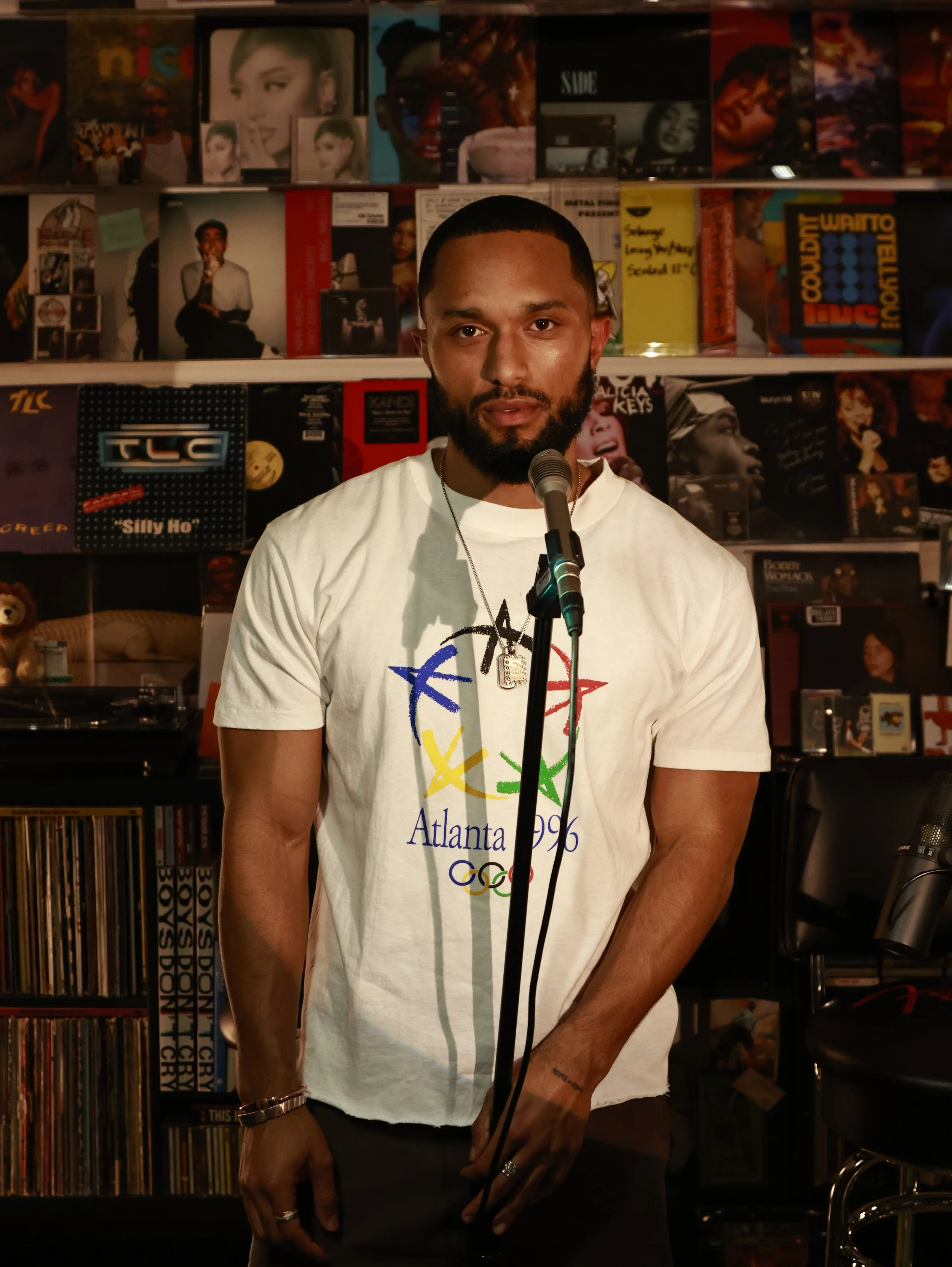 A man with a beard and short hair standing in front of a microphone, wearing a white T-shirt with Atlanta 1996 Olympic logo, in a room filled with music records and posters.