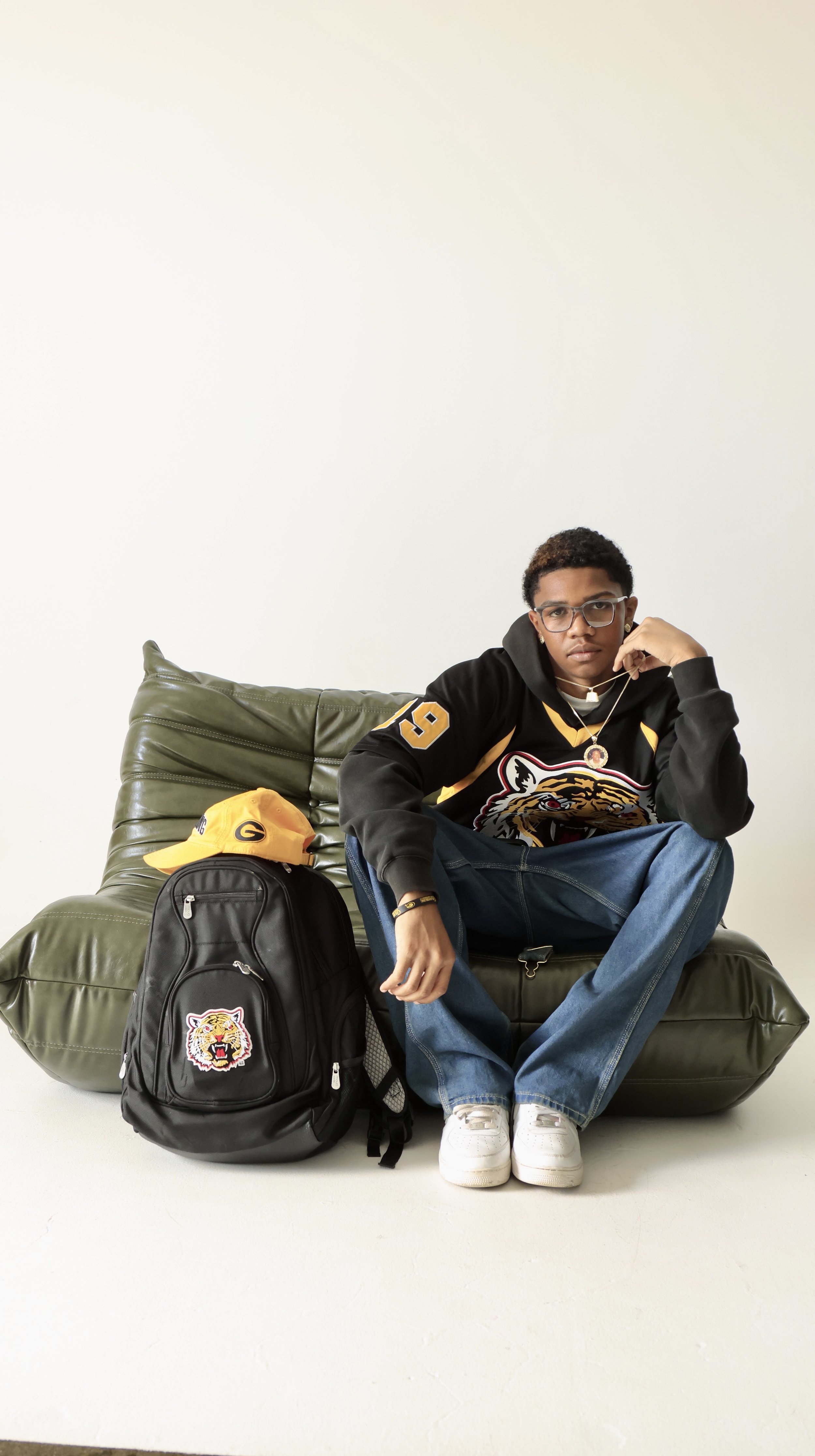 Young man sitting on a green leather couch with a black backpack, yellow cap, and wearing a black hoodie with a tiger logo, jeans, and white sneakers in a studio with a plain white background.