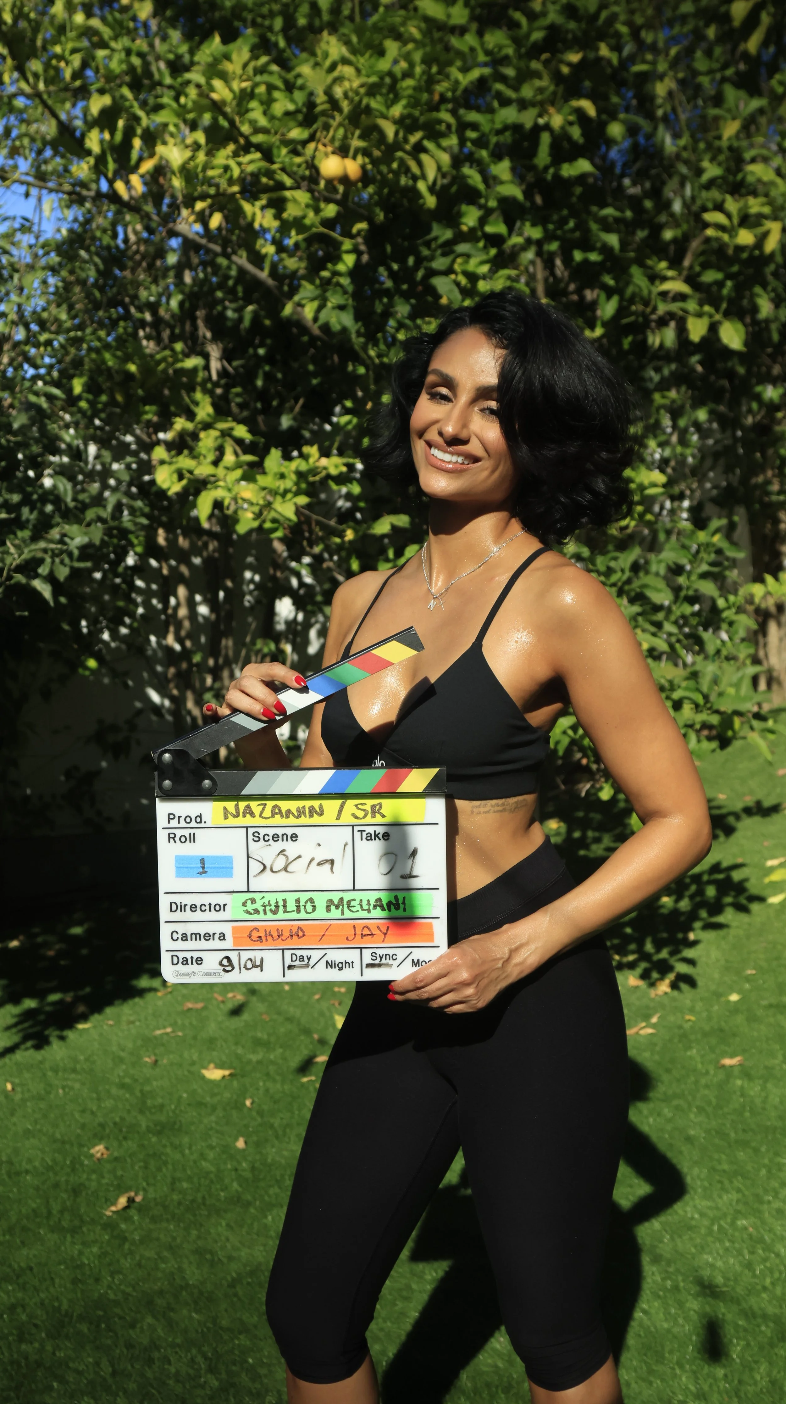 A smiling woman in a black sports bra and black leggings outdoors, holding a film slate, standing in front of greenery and a fruit tree with yellow fruits.