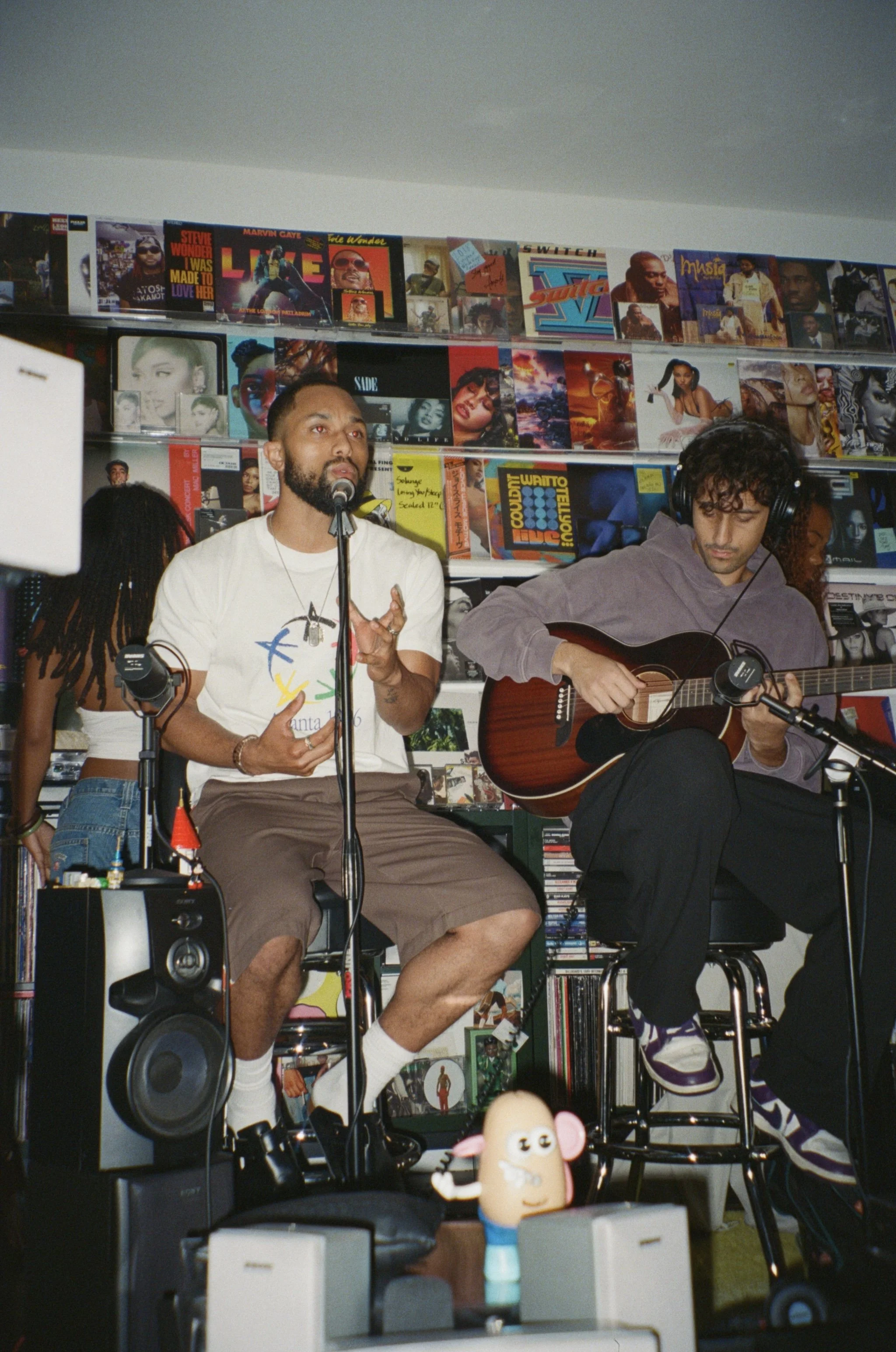 Two men performing music in a record store, one singing into a microphone and the other playing an acoustic guitar, with shelves of vinyl records in the background.