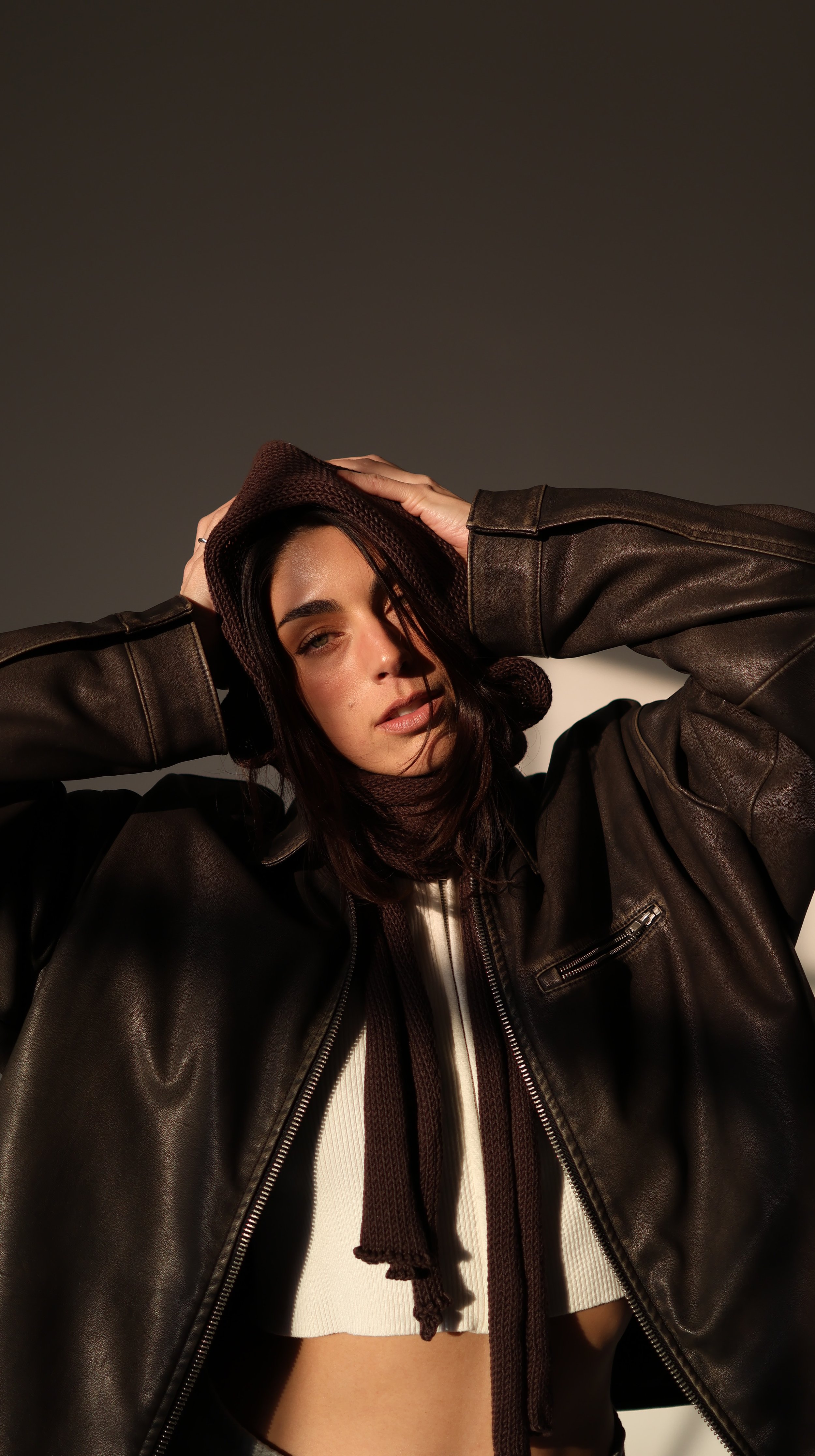 A woman with dark hair wearing a brown hat, a dark leather jacket, and a scarf, posing with her hands on her hat in front of a plain background.