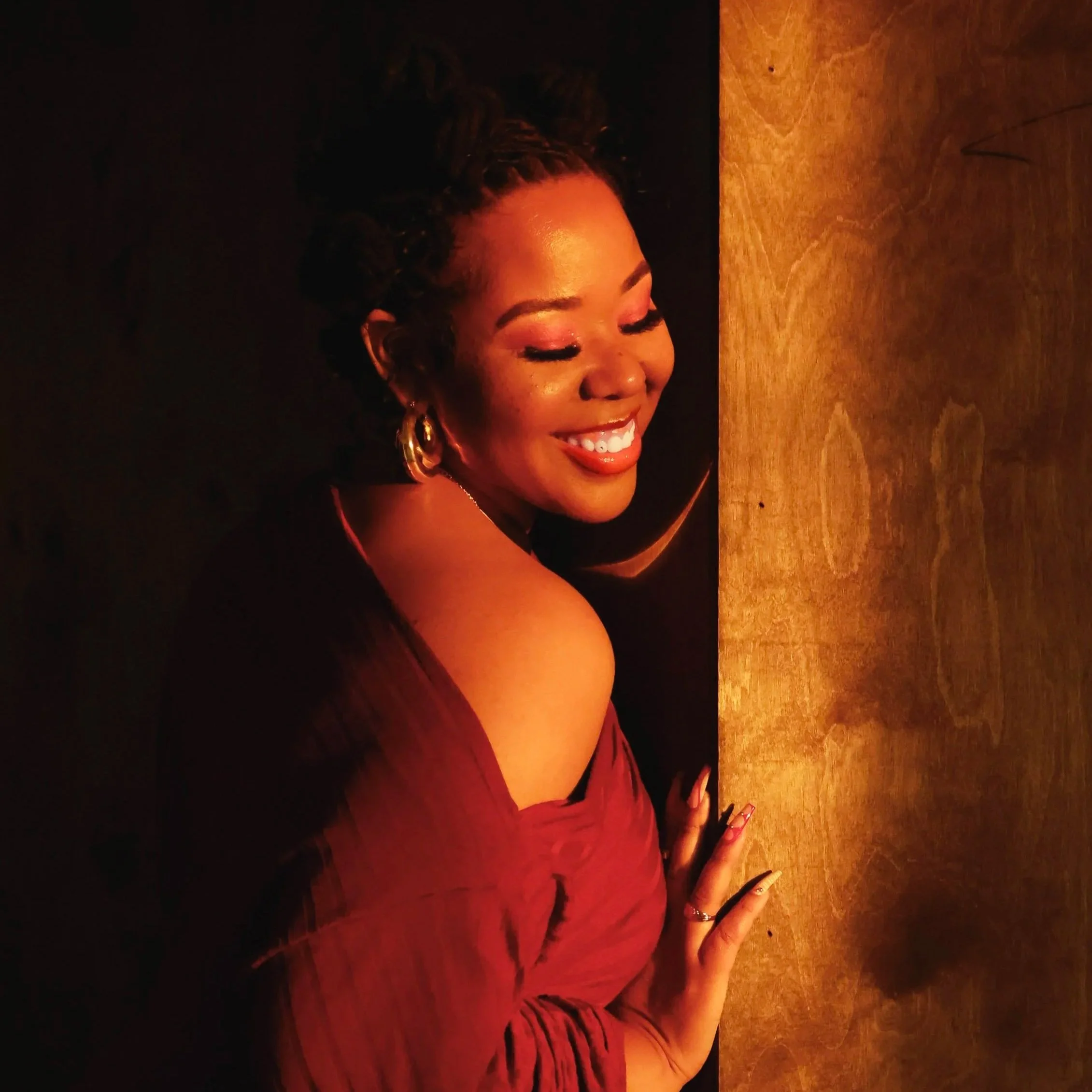 A woman with curly hair, wearing a burgundy off-shoulder dress and gold hoop earrings, smiling with closed eyes and leaning against a wooden surface in a dimly lit environment.