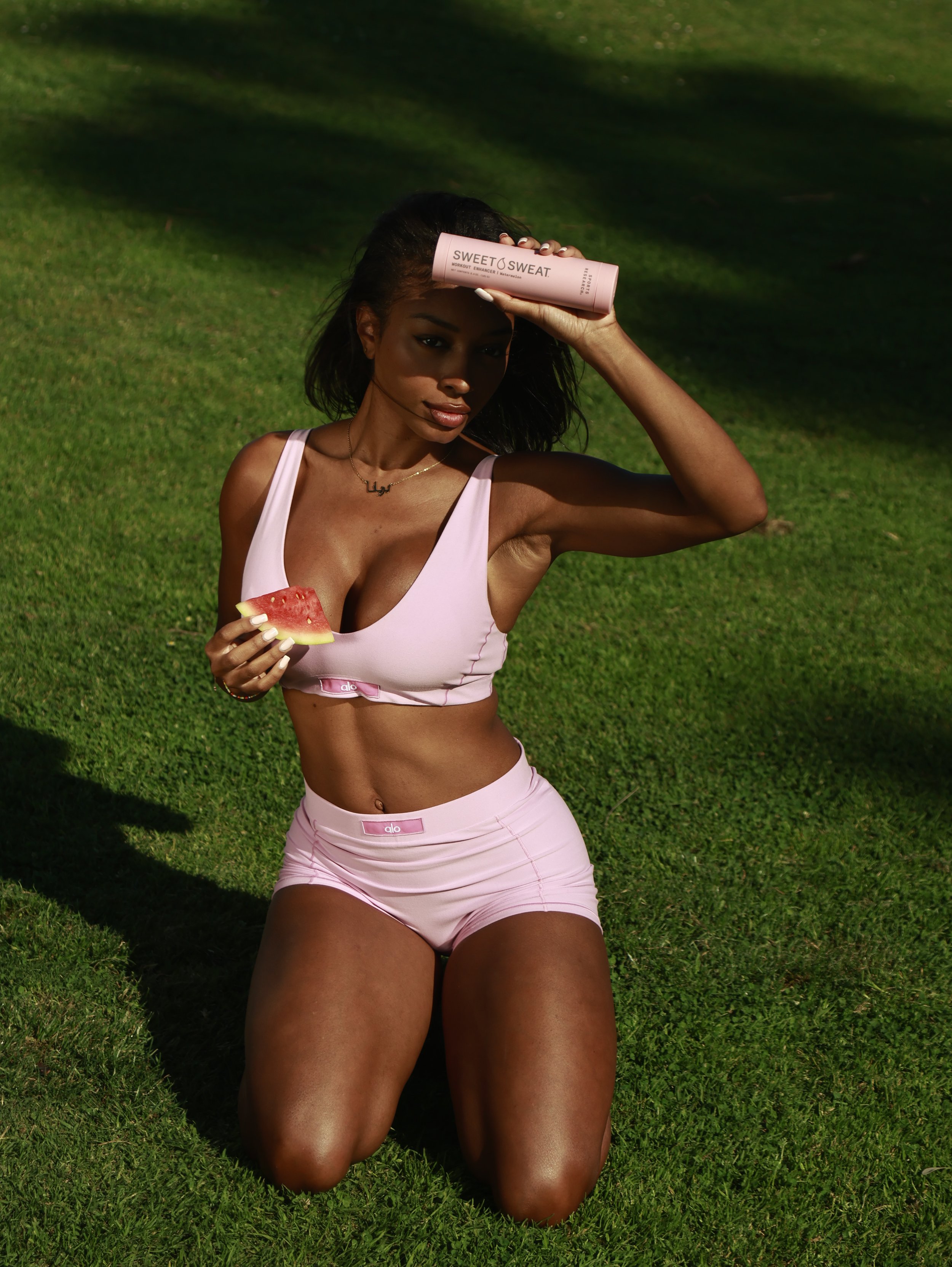 A woman kneeling on grass, holding a slice of watermelon in one hand and a pink cylindrical container labeled "Sweet Sweat" on her forehead with the other hand, wearing a pink sports bra and matching shorts.