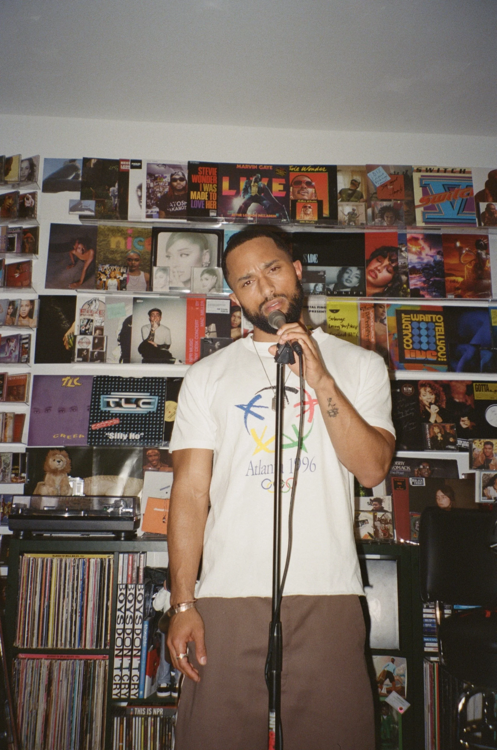 A man holding a microphone in front of a colorful wall of album covers and photographs inside a record store.