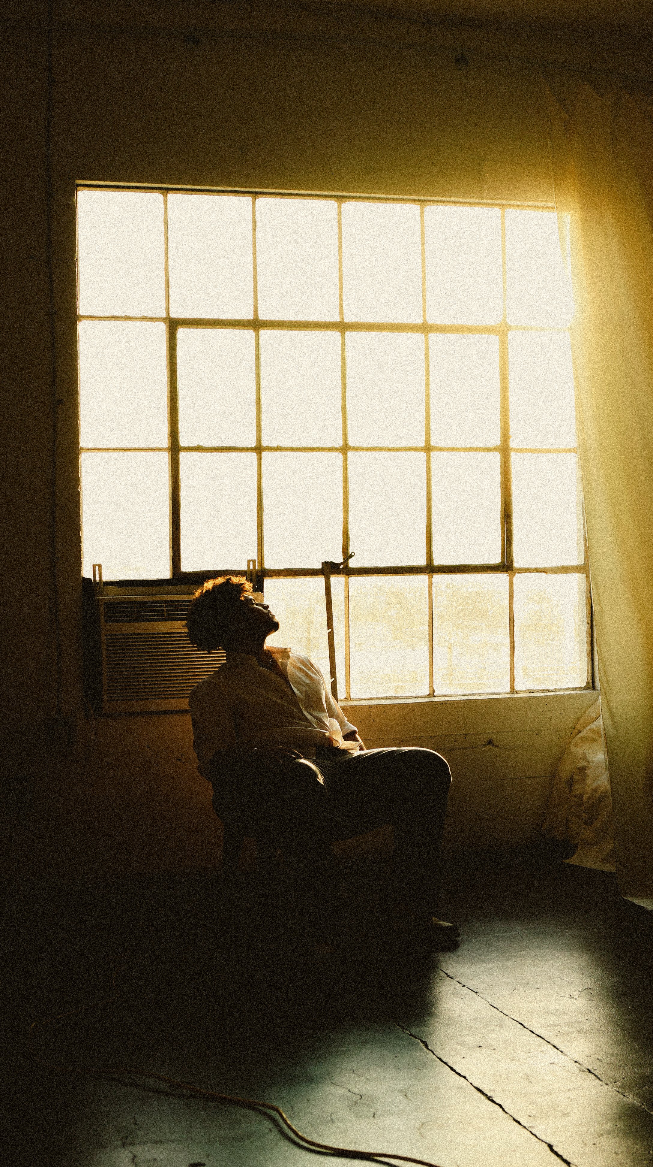 A person sitting on a chair, looking towards a large backlit window with sunlight streaming in, creating a silhouette effect.