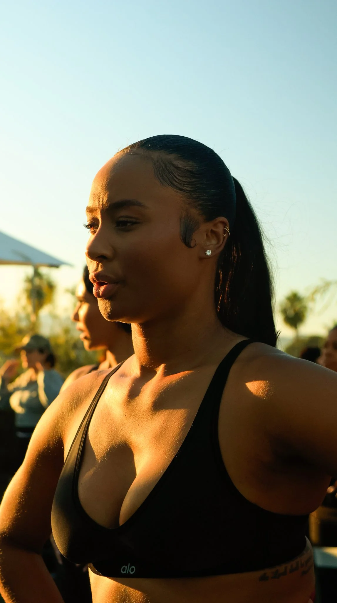 A woman in athletic wear participating in an outdoor event, with sunlight illuminating her face and chest, and other participants visible in the background.