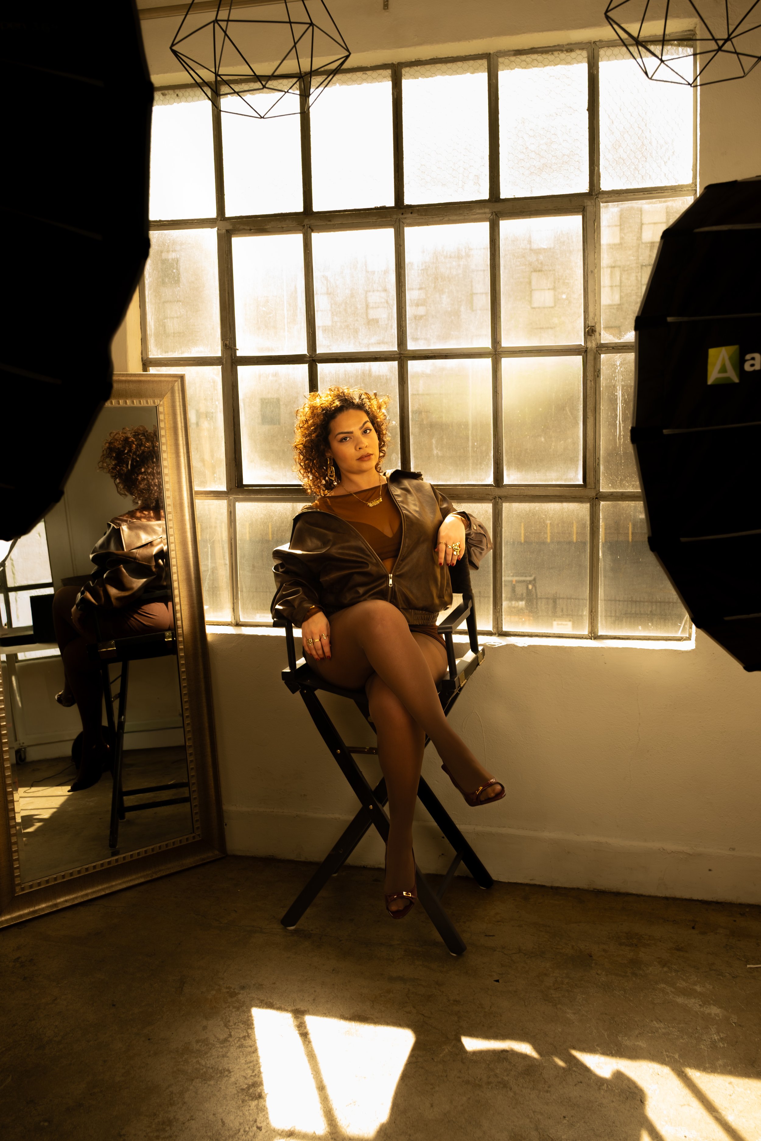 A woman sitting on a director's chair in front of a large industrial-style window with sunlight streaming in, surrounded by photography equipment and a full-length mirror.
