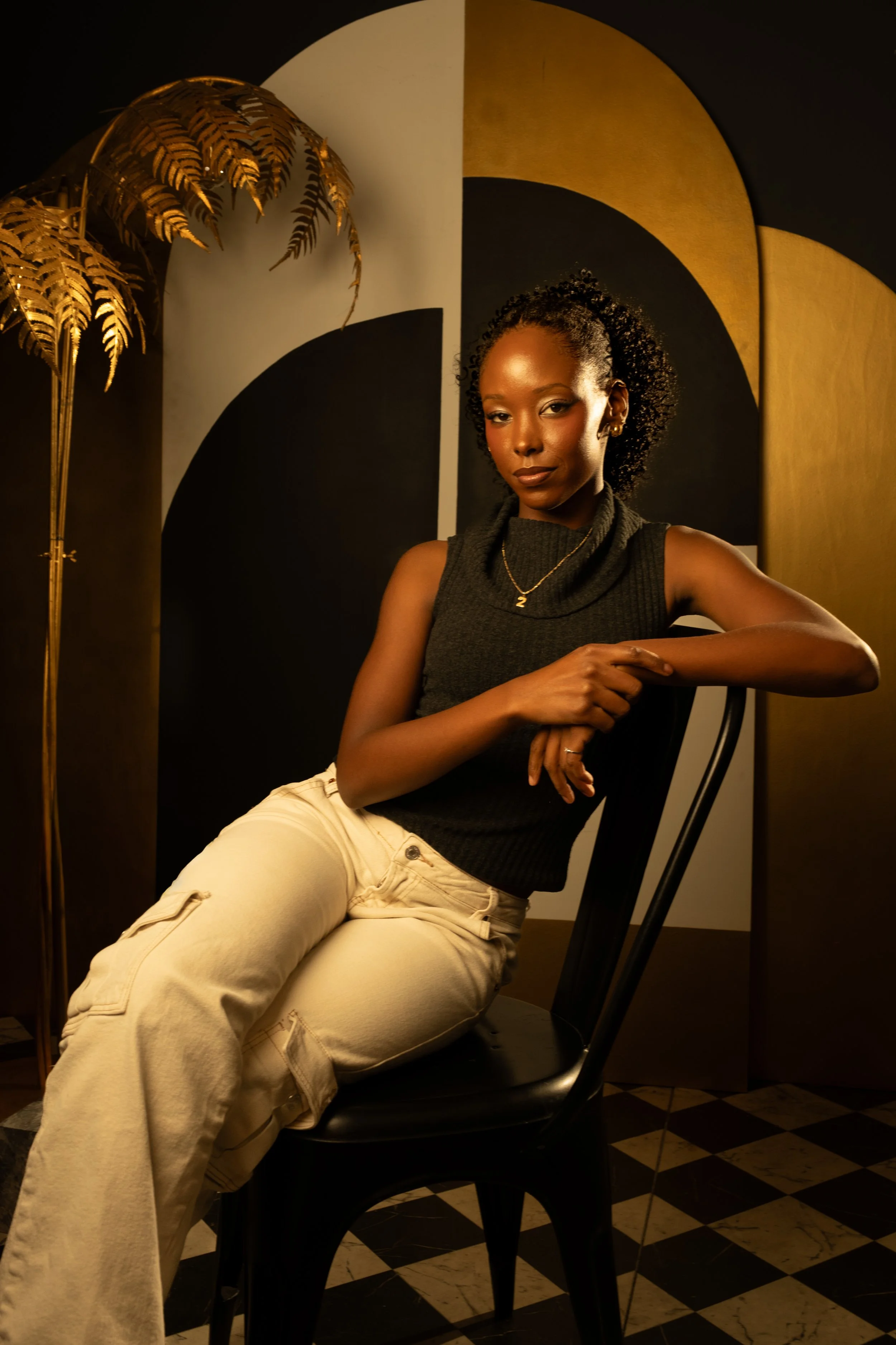 A woman with curly hair sitting on a black chair in a room with black, gold, and white decor, including gold leaves and geometric background panels.