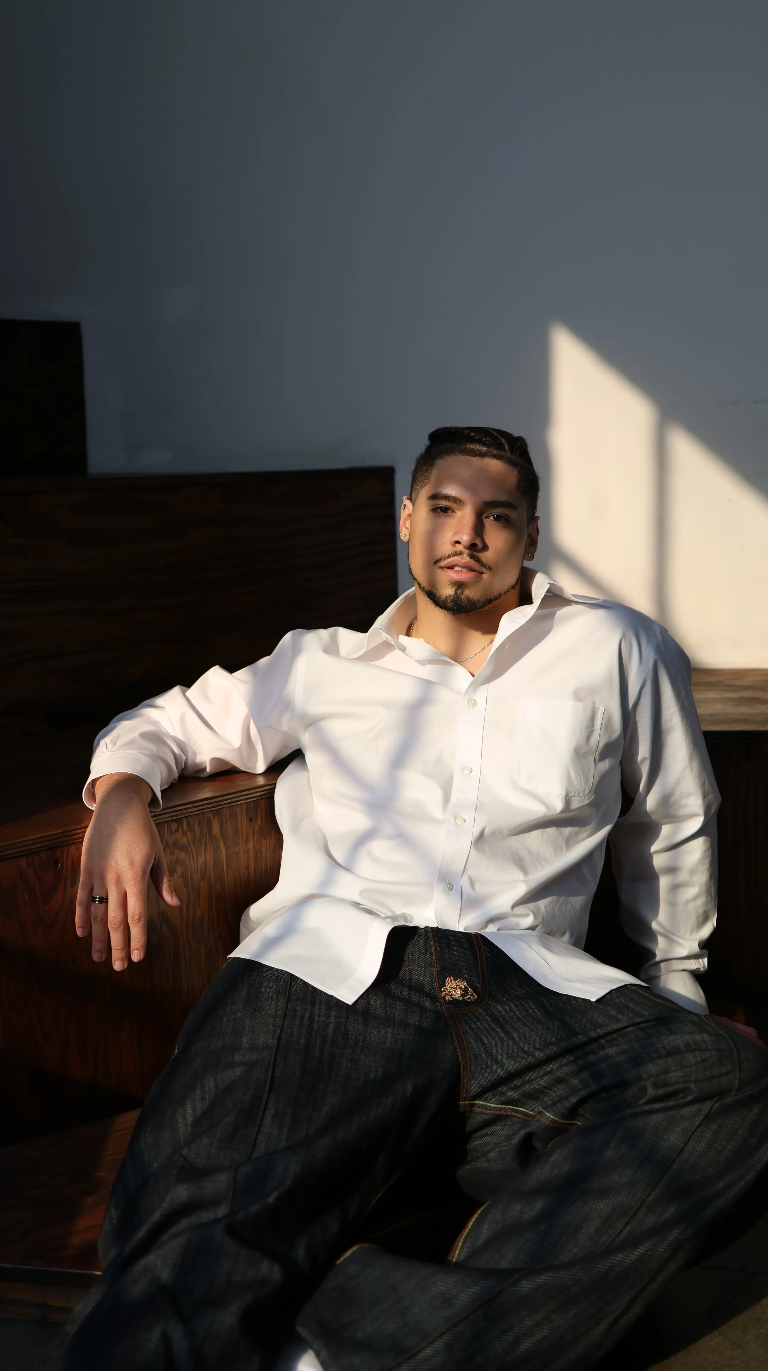 A young man with dark hair, a goatee, wearing a white button-up shirt and dark jeans, sitting on a wooden bench with sunlight creating shadows on the wall behind him.
