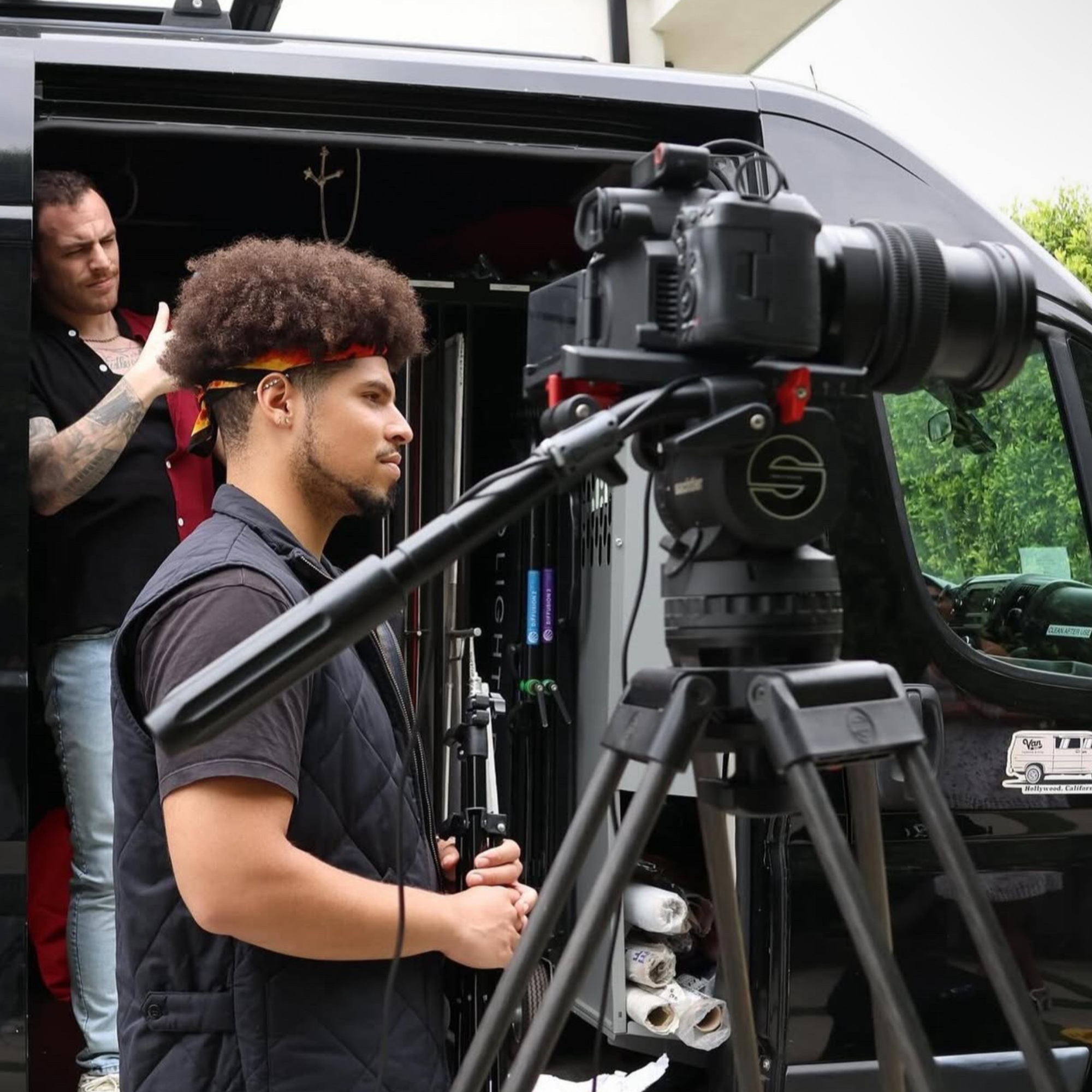 A person with curly hair and a headband, operating a camera mounted on a tripod during a video shoot, with another person in the background adjusting equipment outside a vehicle.