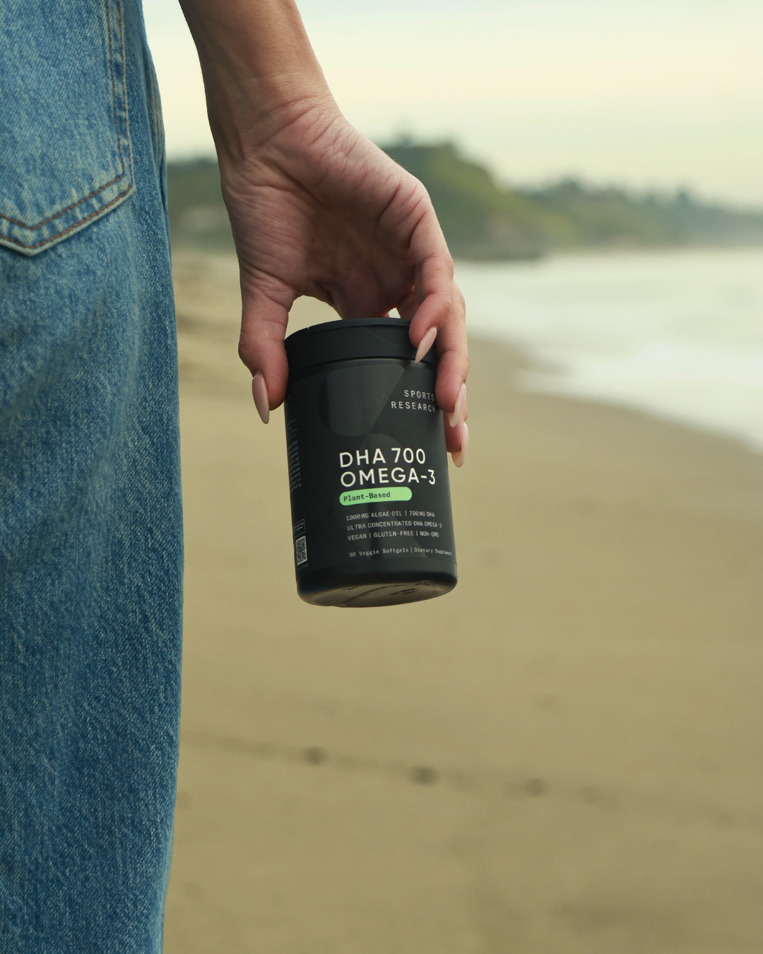 Person holding a bottle of DHA 700 Omega-3 supplement on the beach with blurred shoreline and ocean in background.
