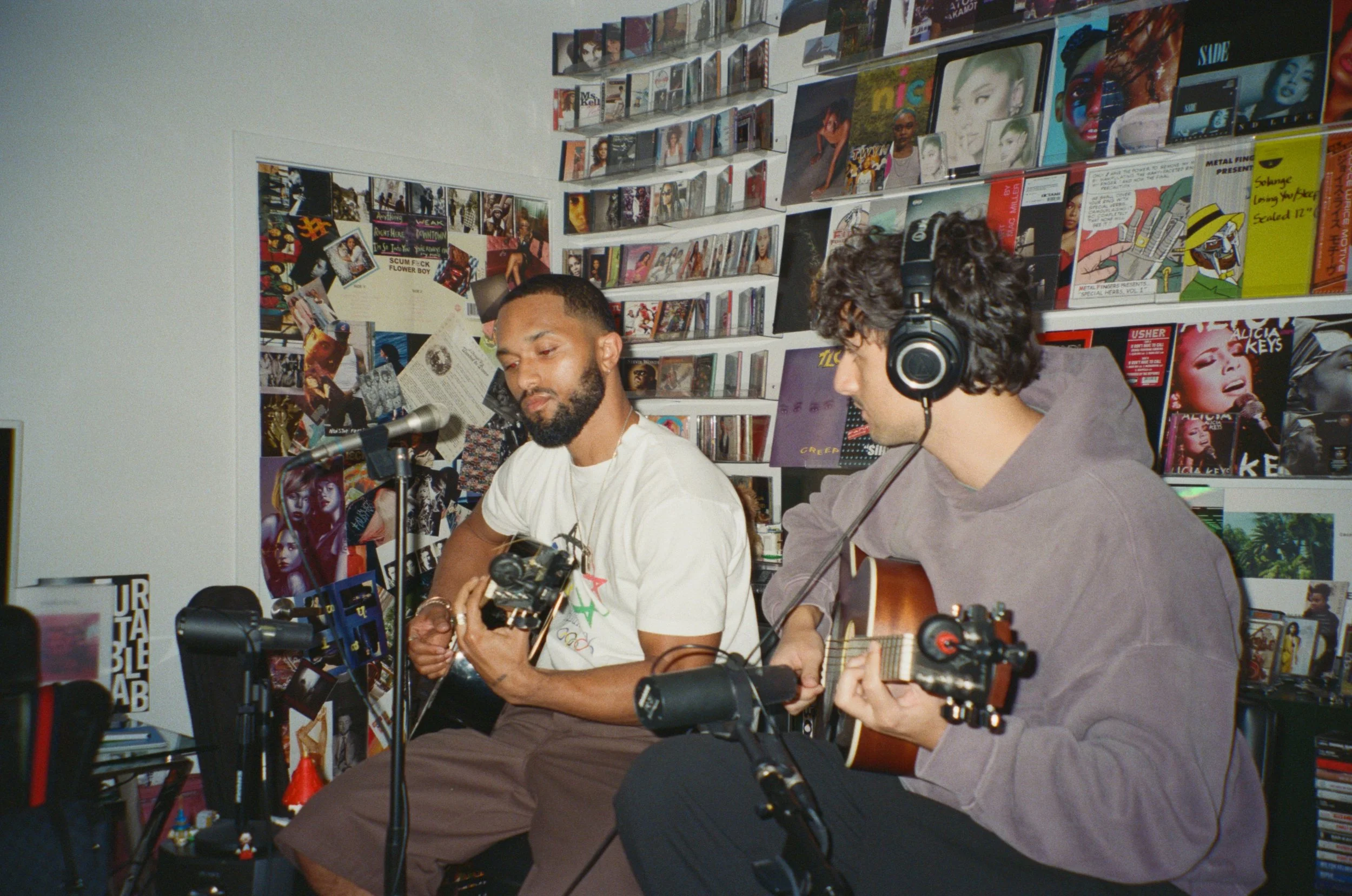 Two men sitting in a music store, one with a guitar and the other with an acoustic guitar, surrounded by shelves of CDs and vinyl records.