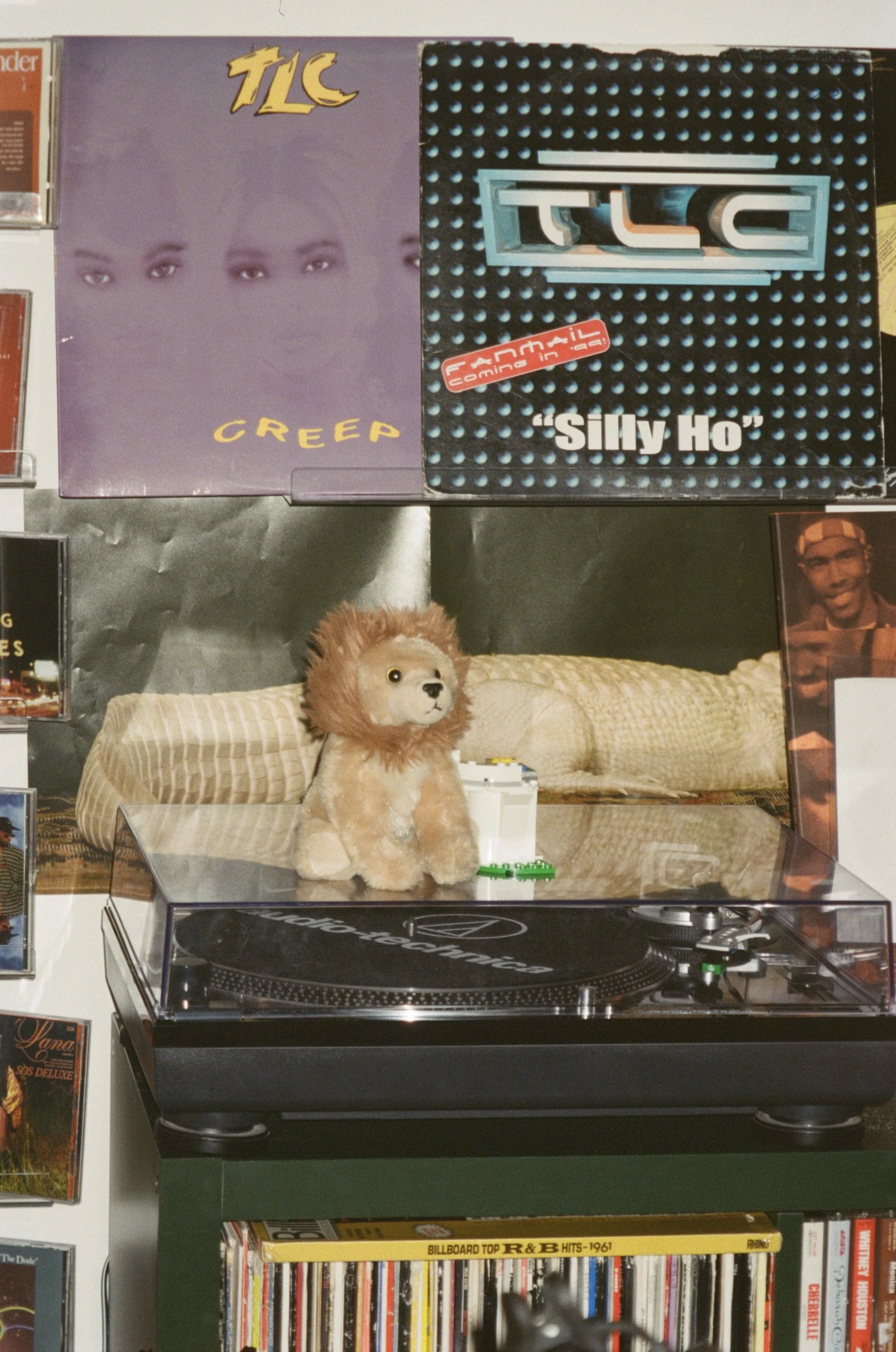 A record player on a green stand, with a plush lion toy and small white Lego-like construction blocks on top. Behind the record player is a brown pillow and a photo of a man. On the wall above, there are album covers, including TLC's 'Creep' and 'Silly Ho'.