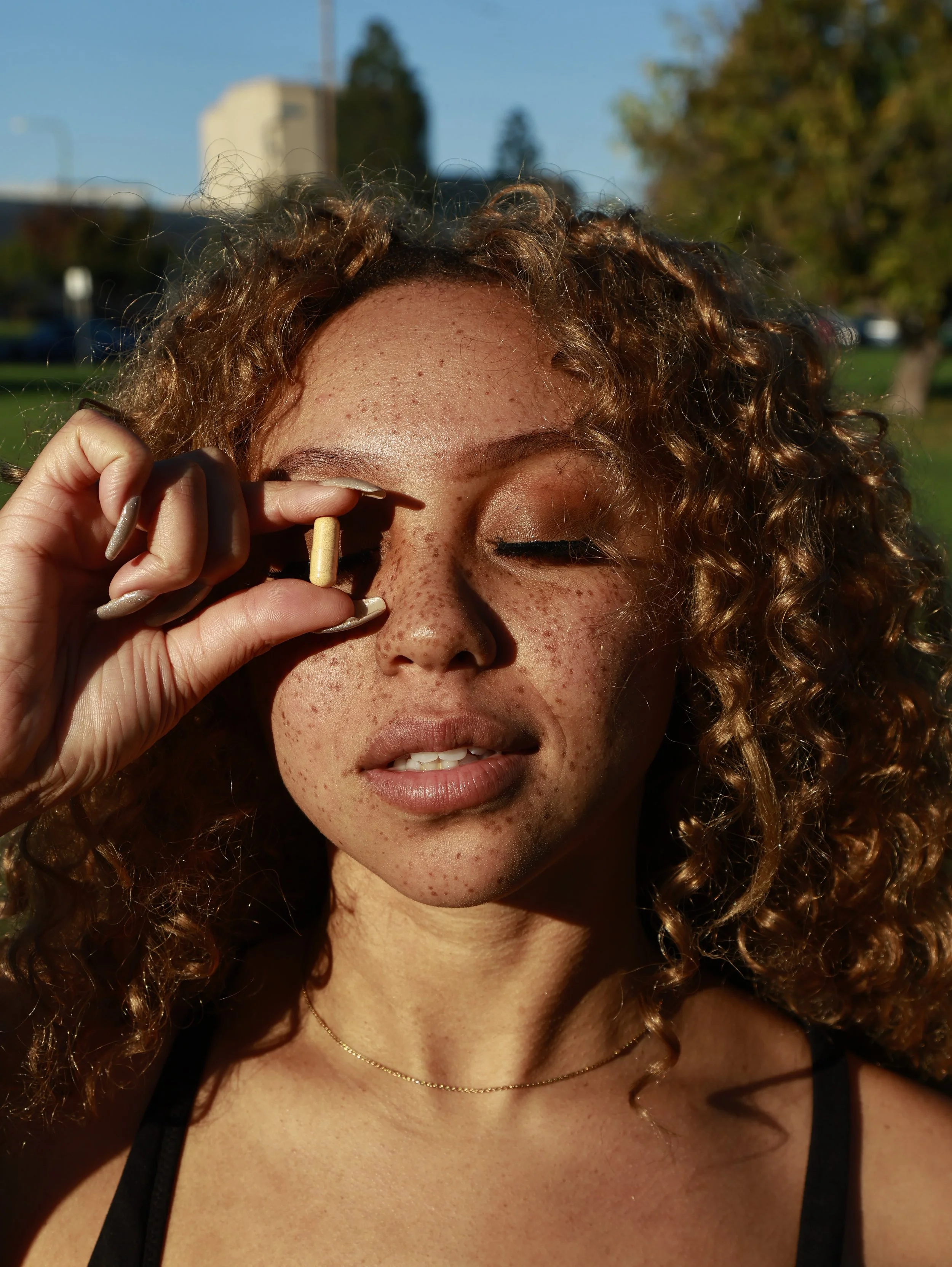 A woman with curly red hair and freckles holding a capsule pill near her eye outdoors during daylight.