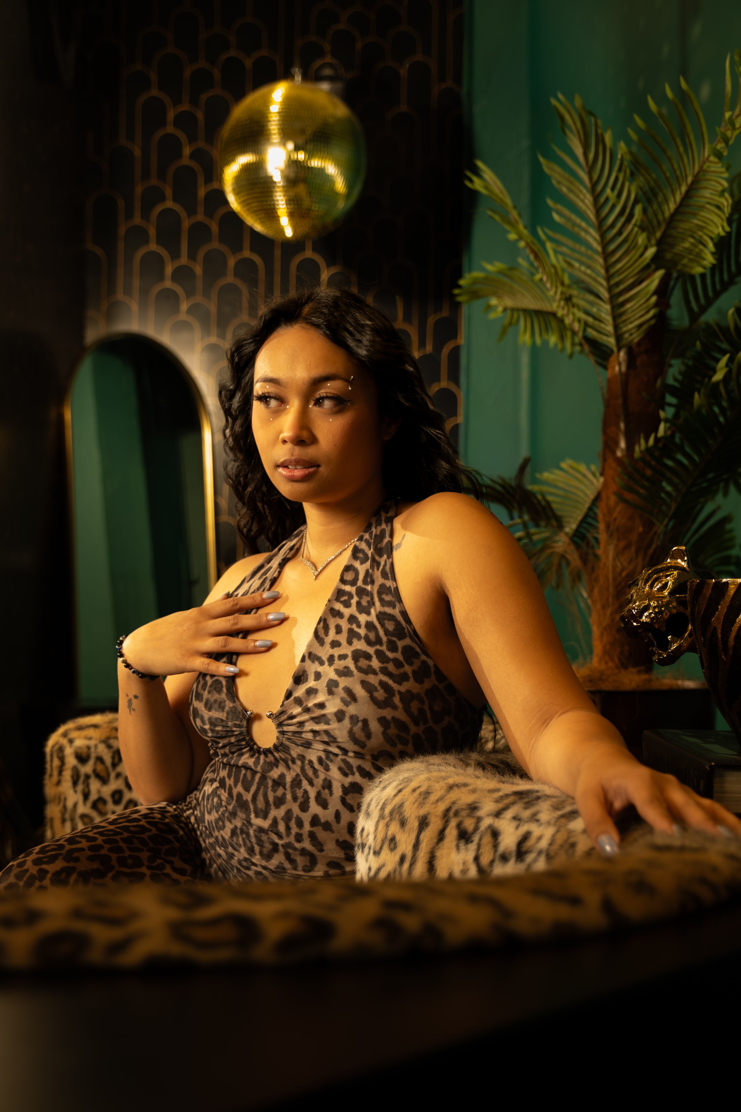 A woman wearing leopard print clothing sitting on a leopard print armchair in a stylish room with green walls, a gold disco ball hanging from the ceiling, a decorative mirror, and a large potted plant.