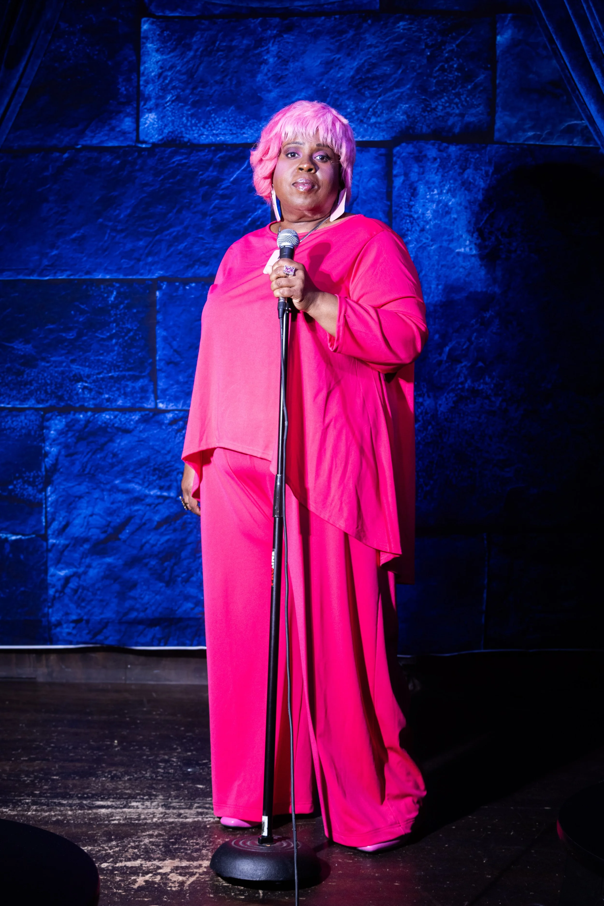 A woman with pink hair, wearing a bright pink outfit, standing on a stage holding a microphone, with a blue textured wall background.