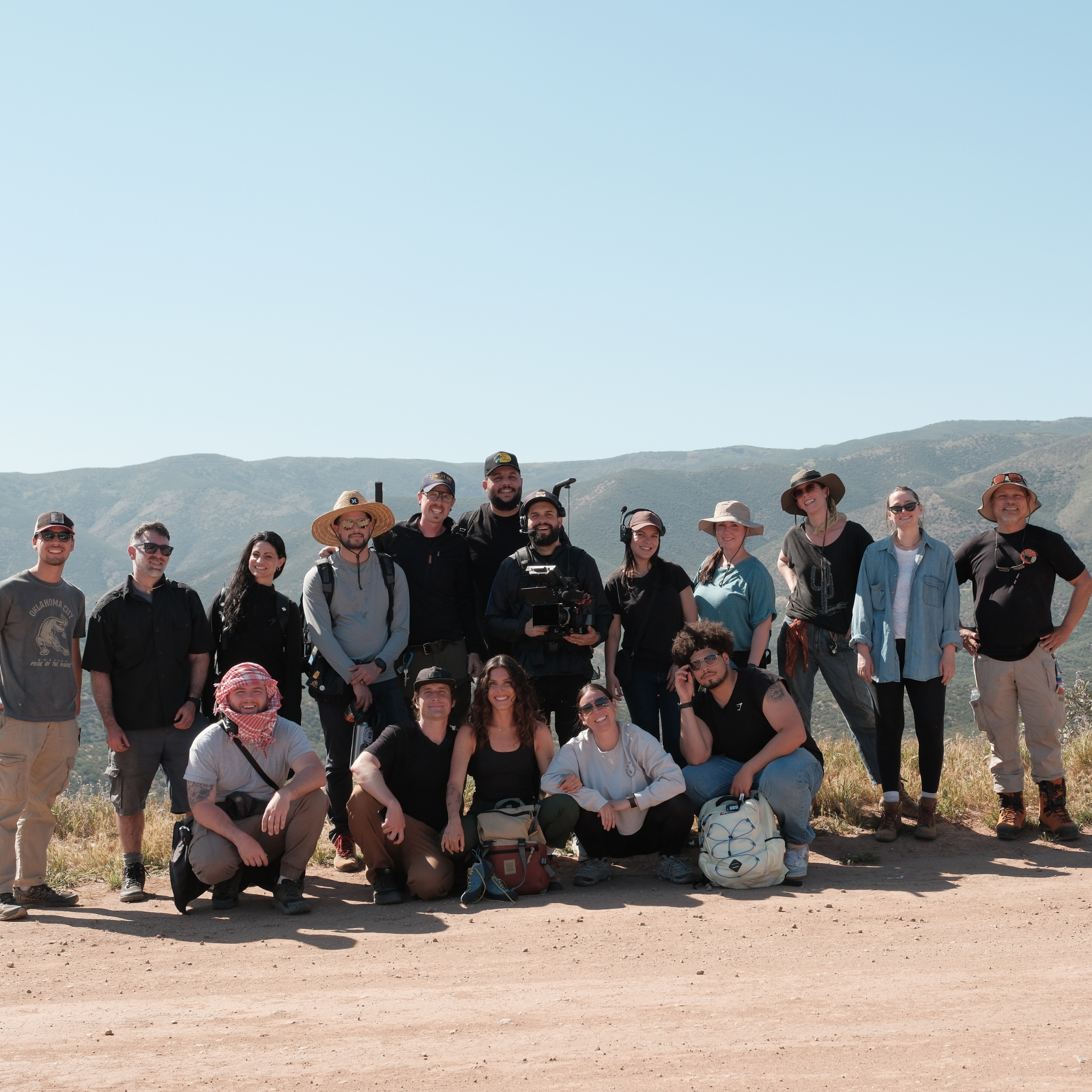 A group of 16 people outdoors on a dirt path with hills and mountains in the background, smiling and posing for a photo.