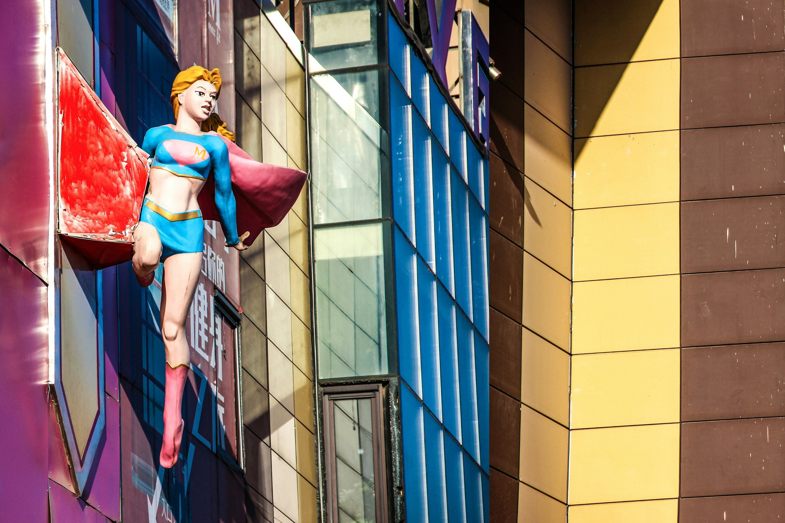 Kunstvolle Statue einer Frau in Superhelden-Kleidung, die aus einem Fenster ausklettert, vor einem modernen Gebäudekomplex mit bunten Fassaden und Glasfenstern.