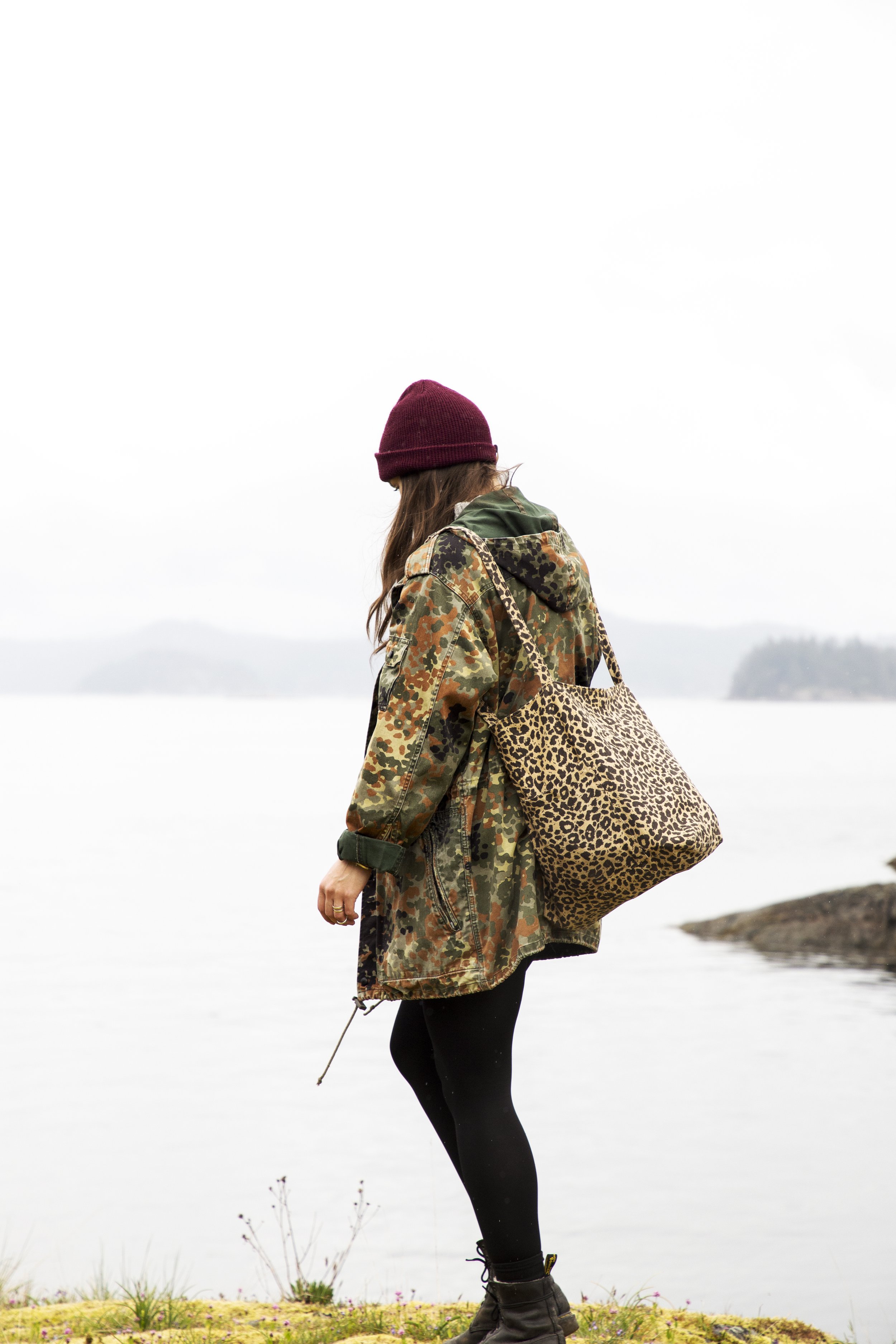 A young woman in a camouflage jacket, black leggings, boots, and a red hat stands by a lake under a cloudy sky, carrying a leopard-print shoulder bag.