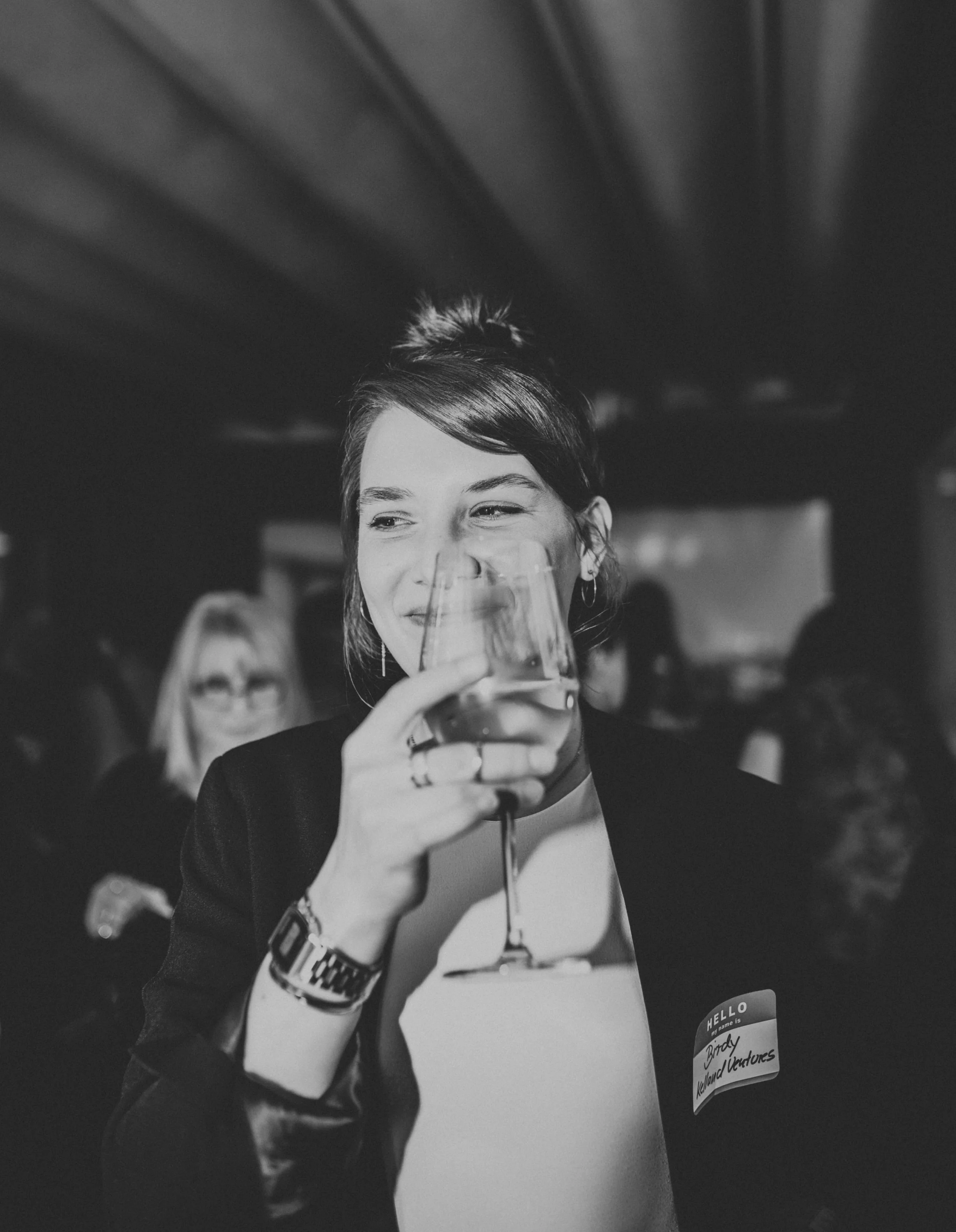 Schwarz-weiß Foto einer lachenden Frau mit dunklen Haaren, die ein Weinglas hält, auf einer Veranstaltung. Sie trägt eine Uhren und hat einen Namensaufkleber mit der Aufschrift 'Hallo, mein Name ist Birdy'.