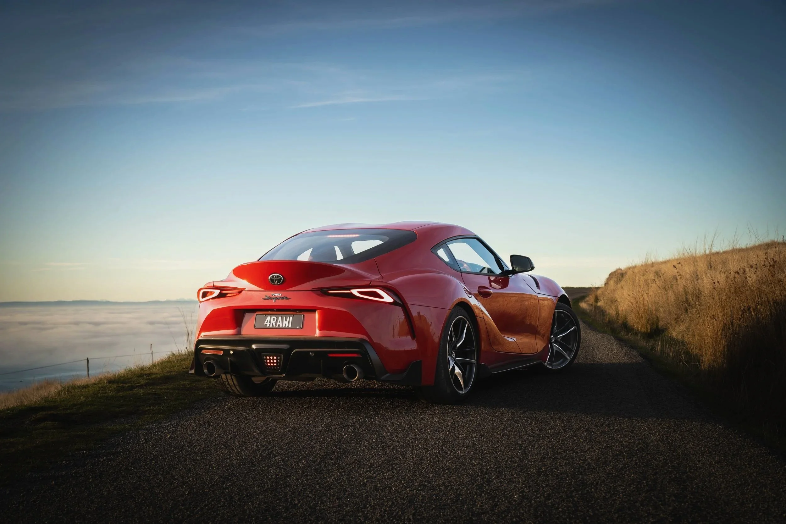 Red sports car parked on a rural road with grass on either side, under a clear sky at sunset or sunrise.