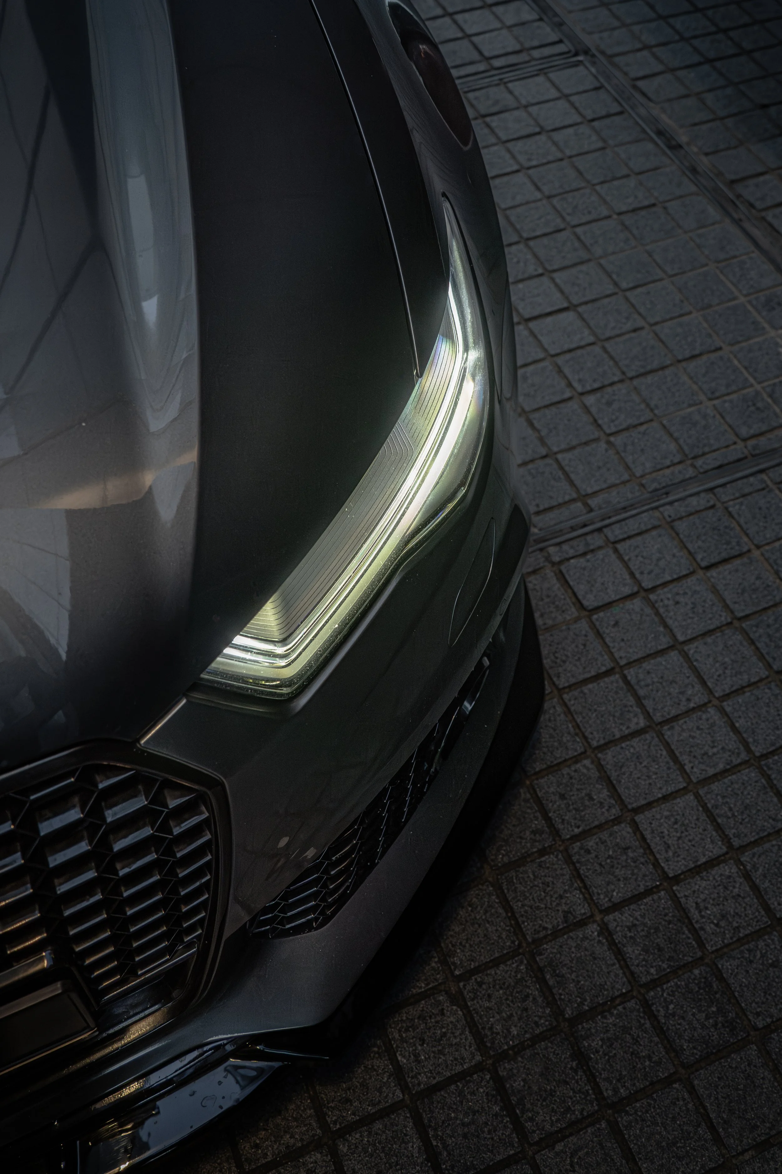 Close-up of a modern black car's front left side, showing the headlight and part of the grille, parked on a brick pavement.