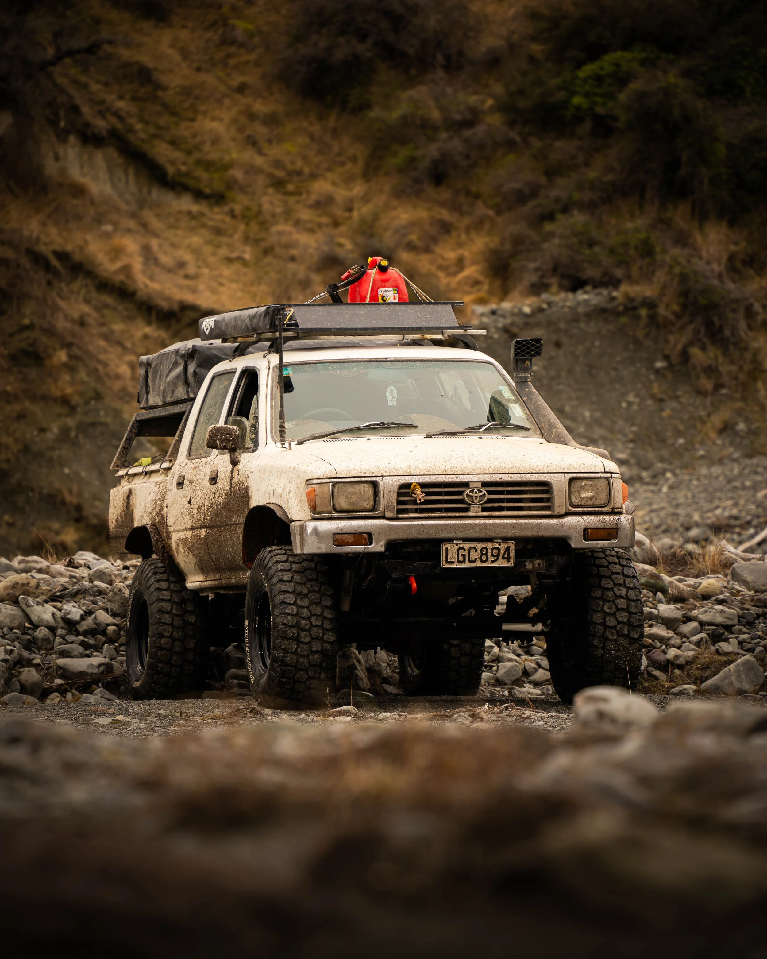 A muddy off-road vehicle, possibly a Toyota, parked on rocky terrain with a mountainous background. The vehicle has large tires and equipment on the roof, including a red fuel container and a fire extinguisher.