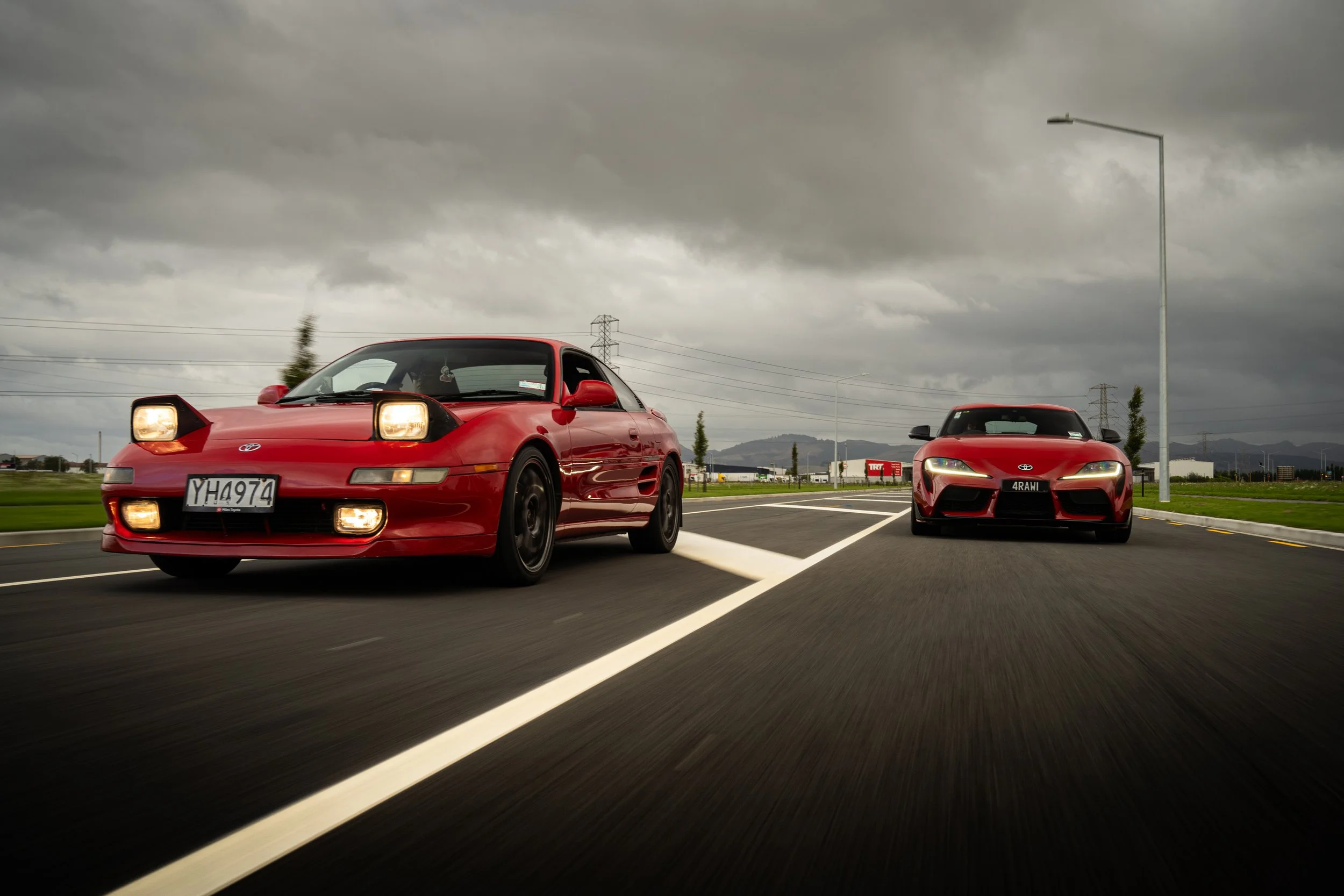 Two red sports cars driving on a cloudy day, one with its pop-up headlights up and the other with headlights on, on a multi-lane road with green fields and mountains in the background.