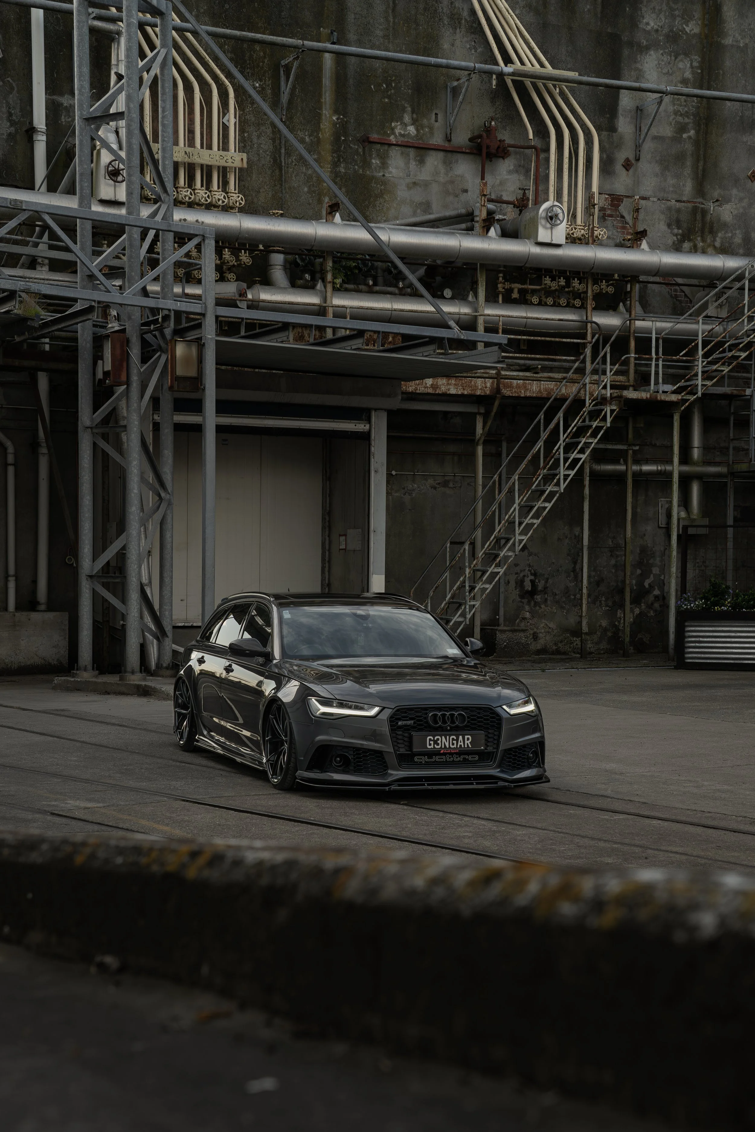 A black Audi sedan parked on an urban street with industrial buildings and metal staircases in the background.
